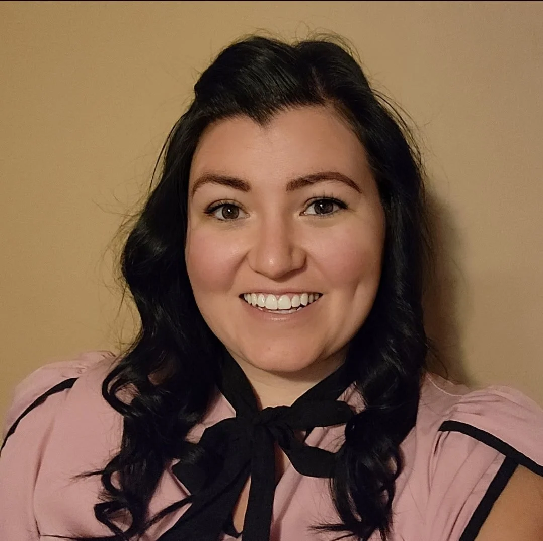 A woman with black wavy hair, smiling, wearing a pink top with black accents and a black ribbon tied at her neck, standing against a beige wall.