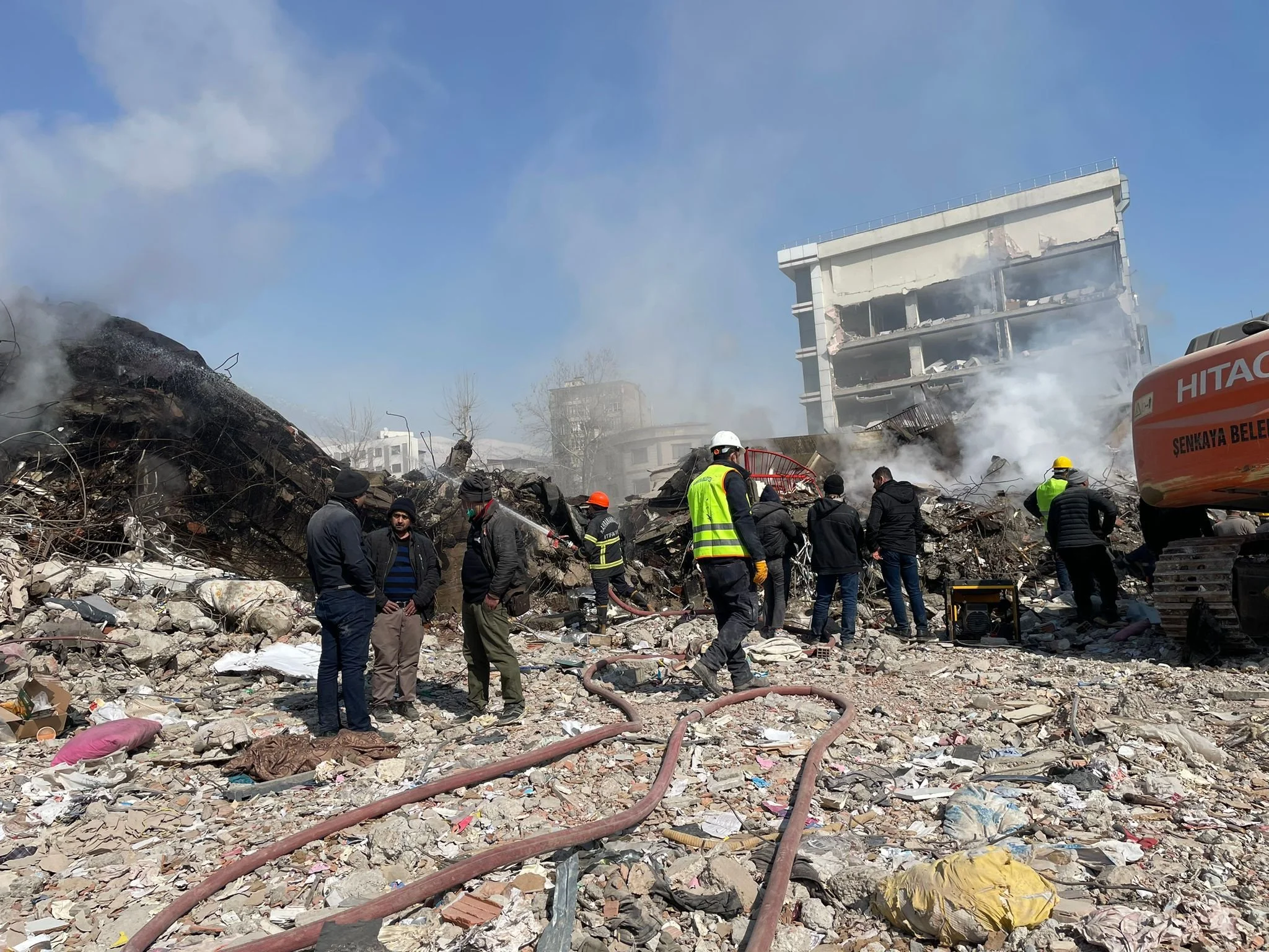 Group of people, including construction workers and emergency responders, standing amidst rubble and debris from a collapsed building, with smoke or dust in the air and a partially damaged building in the background.