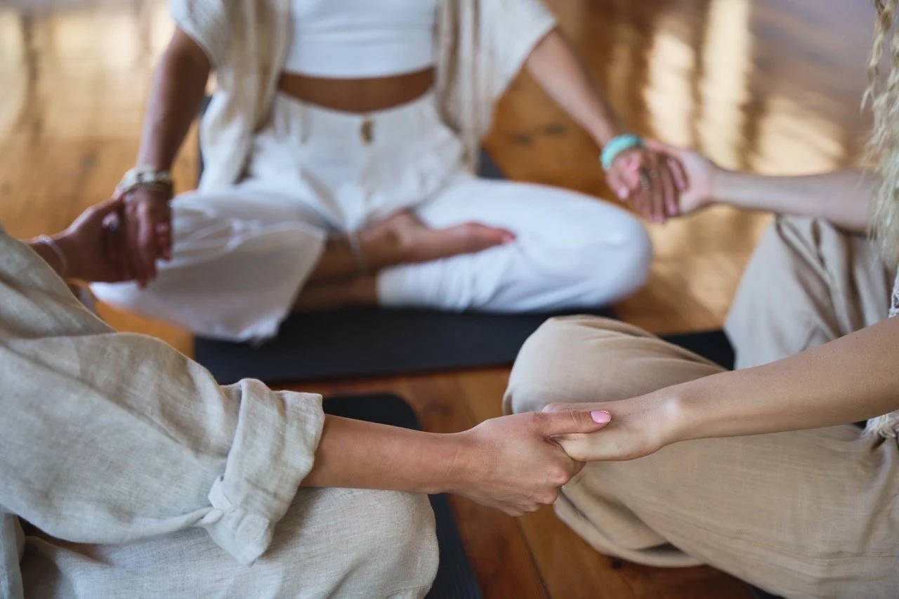 Two people holding hands during a meditation or yoga session with a third person sitting cross-legged in the background.