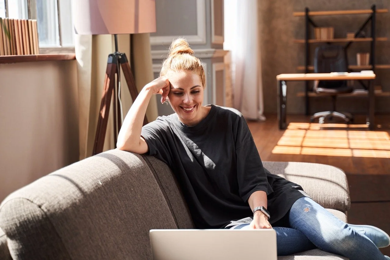 A woman sitting on a gray couch using a laptop, smiling, in a well-lit room with wooden flooring and furniture.