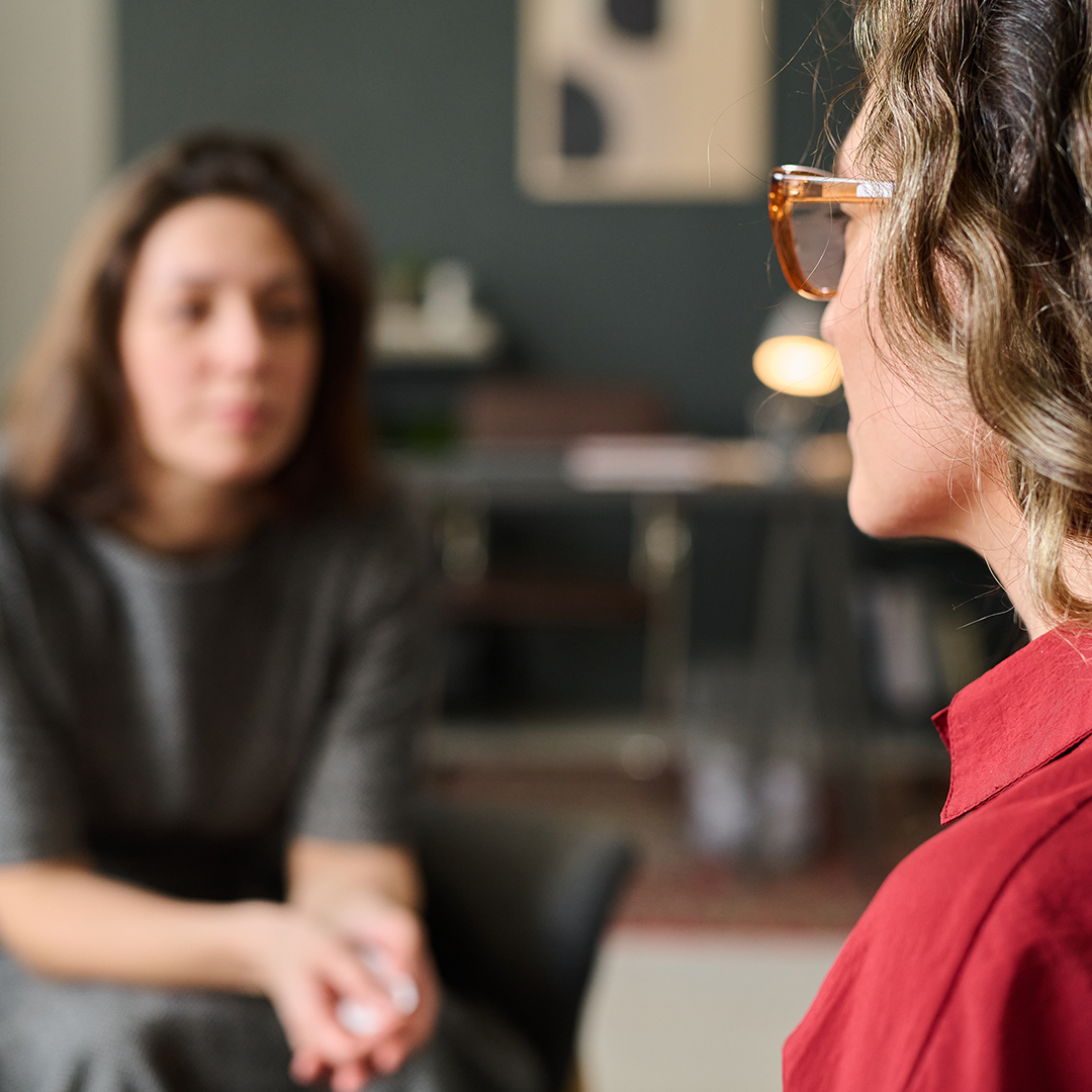 Two women sit across from each other in a therapy session. The woman in the foreground has wavy hair, glasses, and wears a red shirt. The woman in the background is slightly blurred, has straight hair, and wears a dark patterned top.