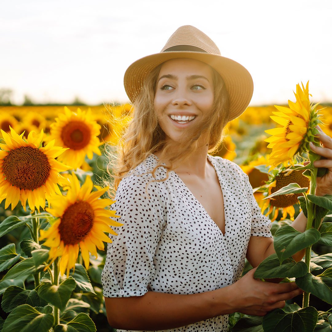 A woman smiling and holding a sunflower in a sunflower field during daytime, wearing a sun hat and a white dress with black polka dots.