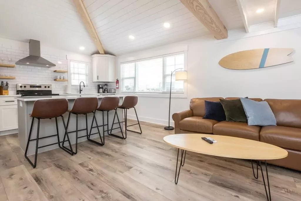 Modern open-concept living room and kitchen with white walls, wooden accents, brown leather sofa, wooden coffee table, and kitchen island with four brown chairs.