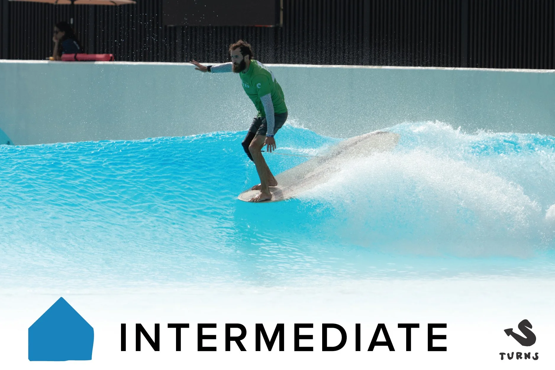 A woman surfing on an indoor wave pool, wearing a green shirt and blue shorts, with a sign indicating 'Intermediate' level at the top.