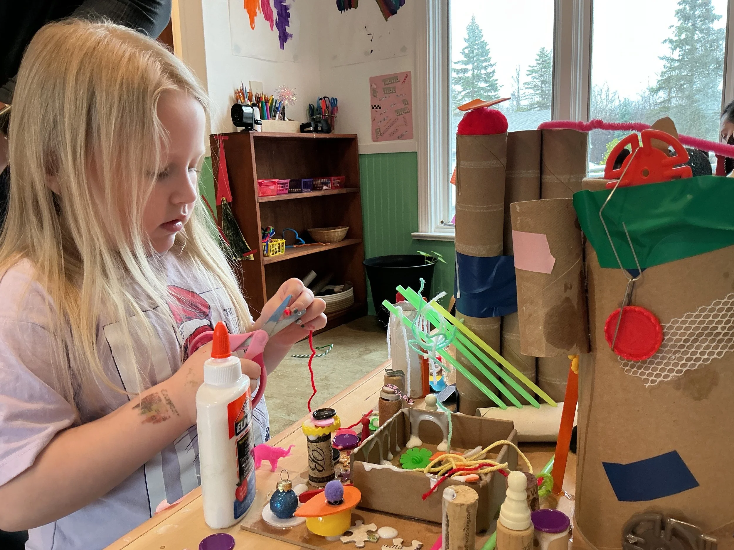 A young girl with long blonde hair and a temporary tattoo on her left arm, concentrating on her crafting project with glue and scissors on a table filled with various craft supplies and small handmade items.