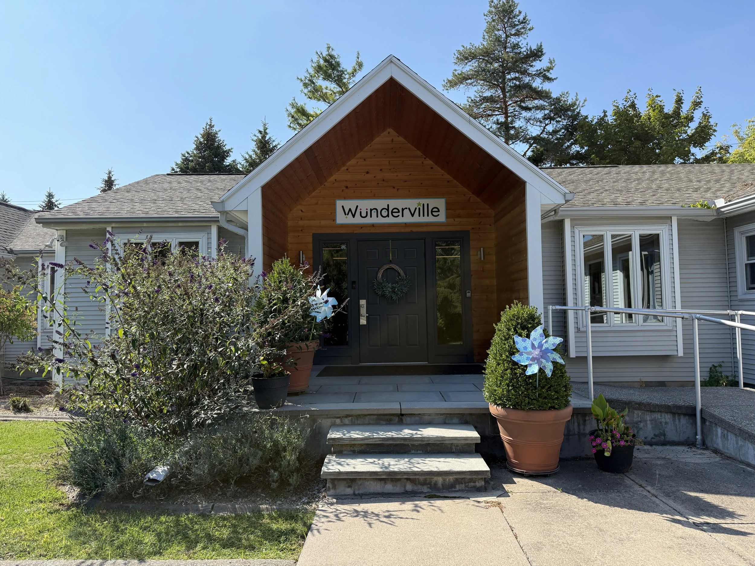 Front of a house with a small porch, stairs, potted plants, greenery, and a sign that reads 'Wunderville' above the door.