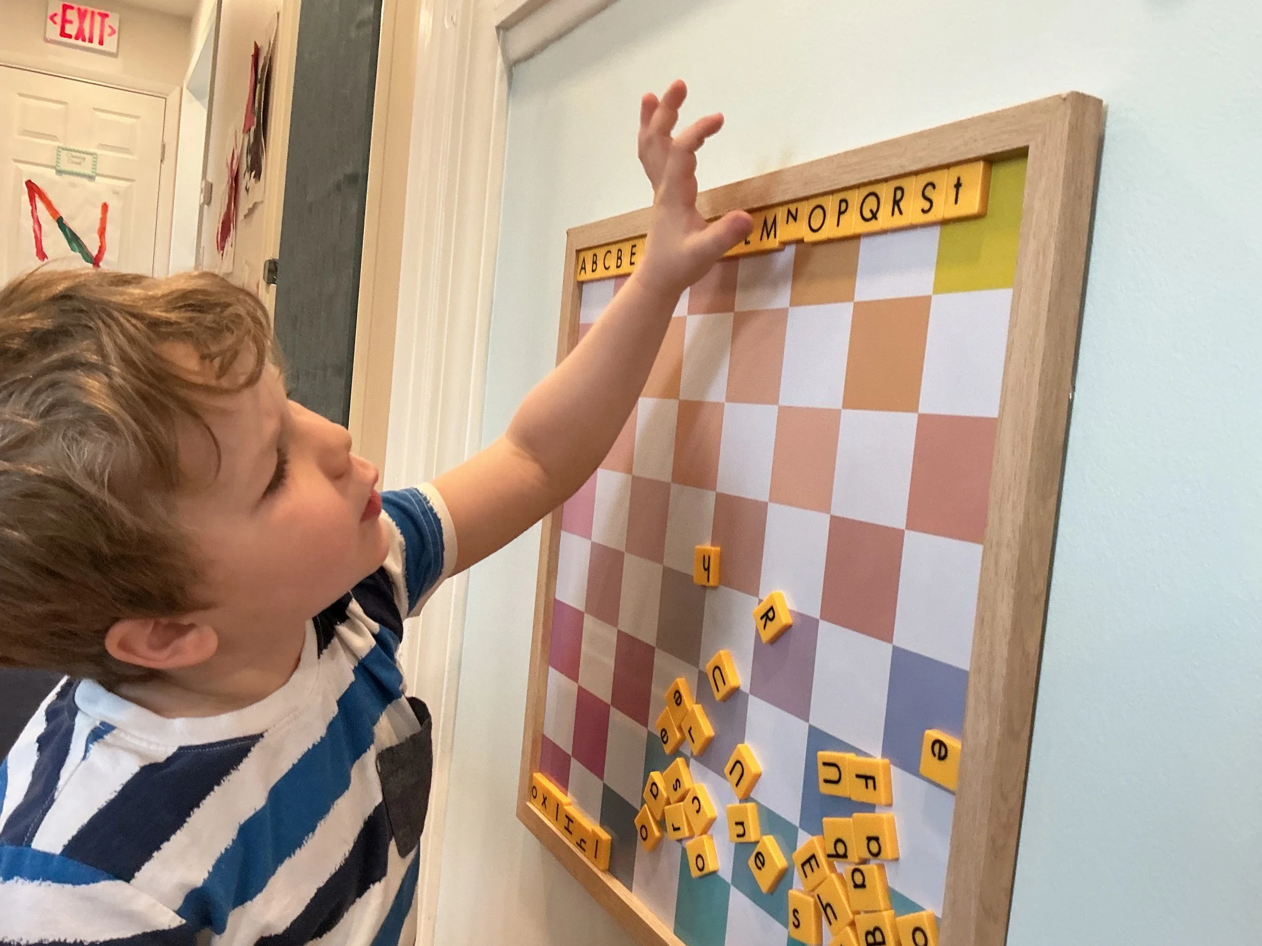A young boy playing Scrabble on a wooden board mounted on a wall, with many letter tiles on the board and some on the surface below.