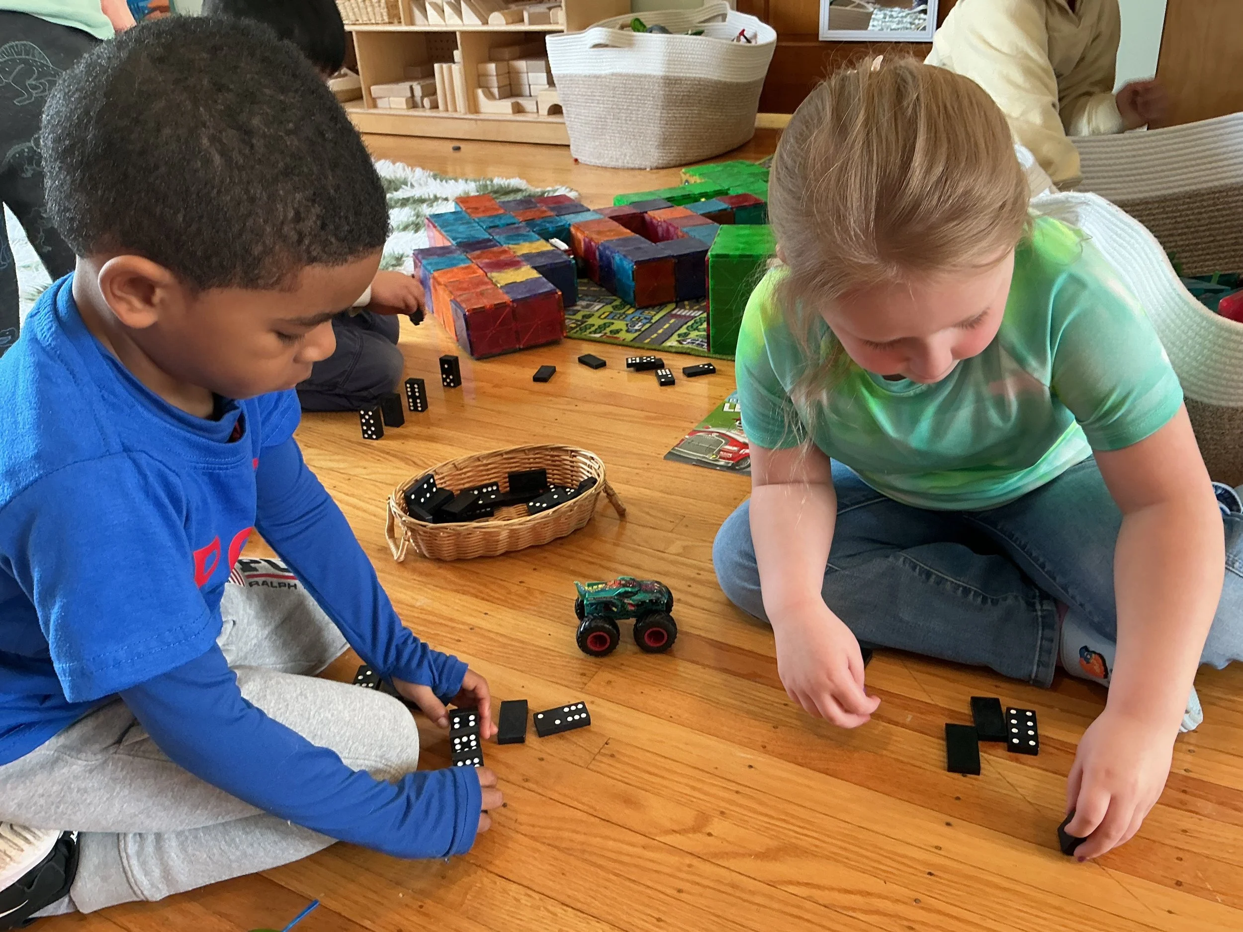 Two children sitting on a wooden floor playing with dominoes and a toy monster truck. There are colorful blocks and dominoes in the background.