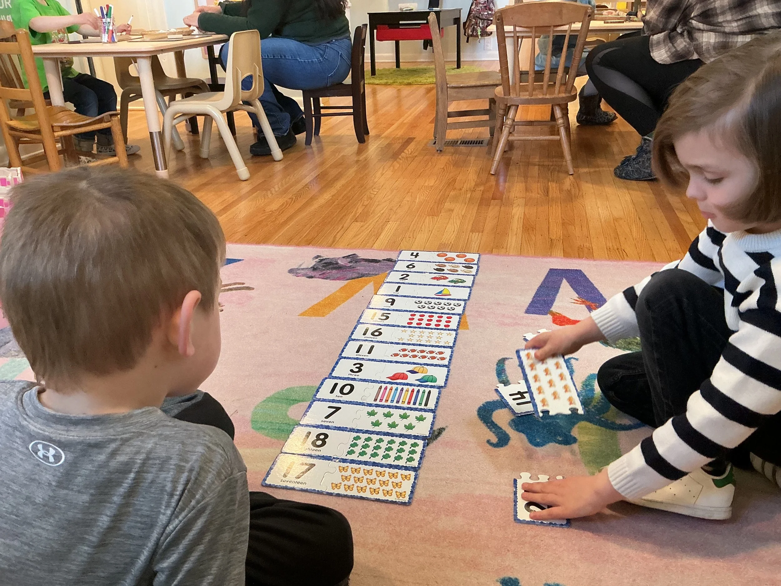 Two children sitting on the floor playing a number and counting game with large, colorful cards laid out in a line. The girl is placing a card while the boy watches.