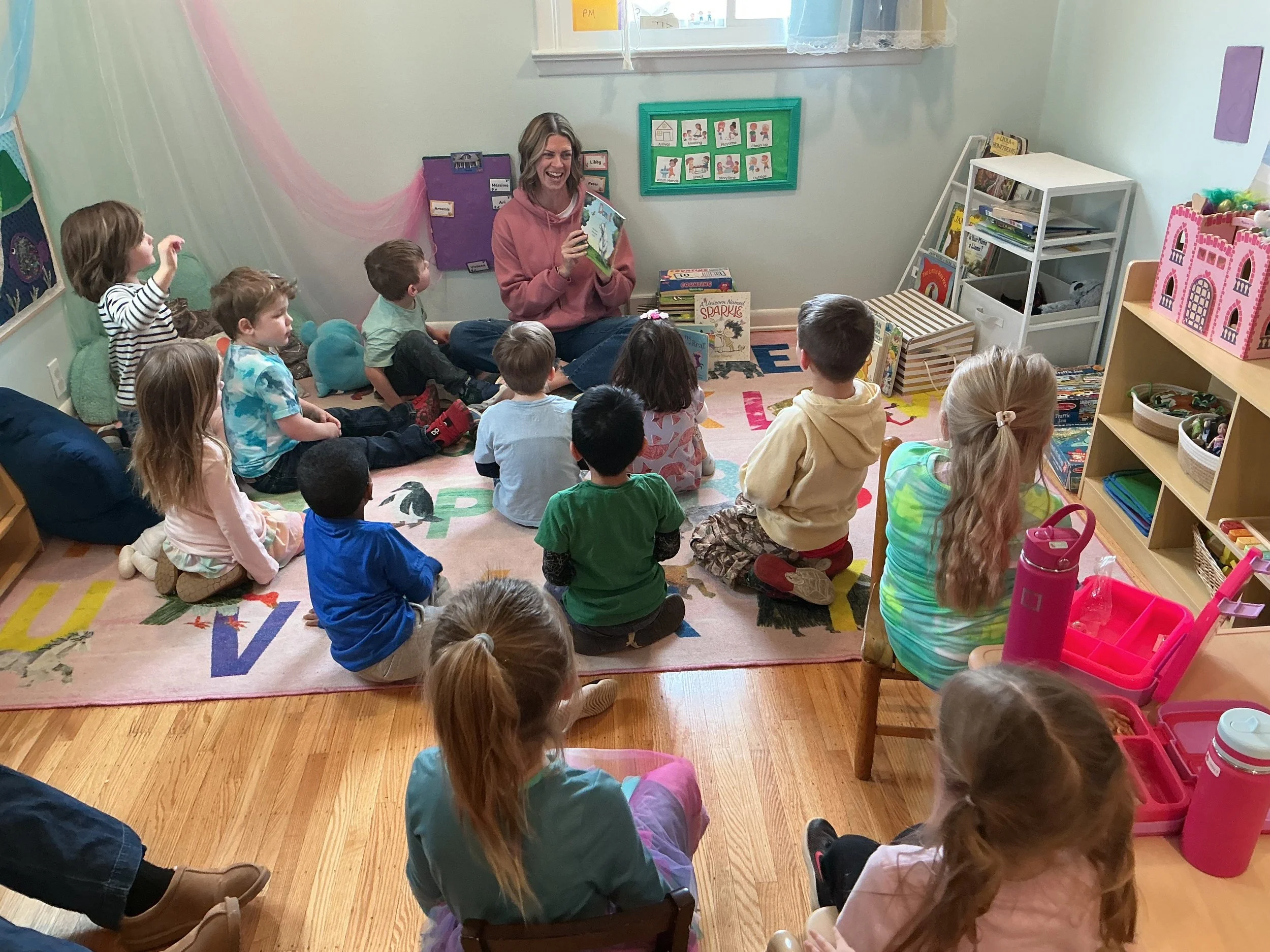 A group of young children sitting on a colorful educational rug in a classroom, listening to a woman reading a book aloud. The room has shelves with books and educational materials, and the children are attentively looking at the woman.