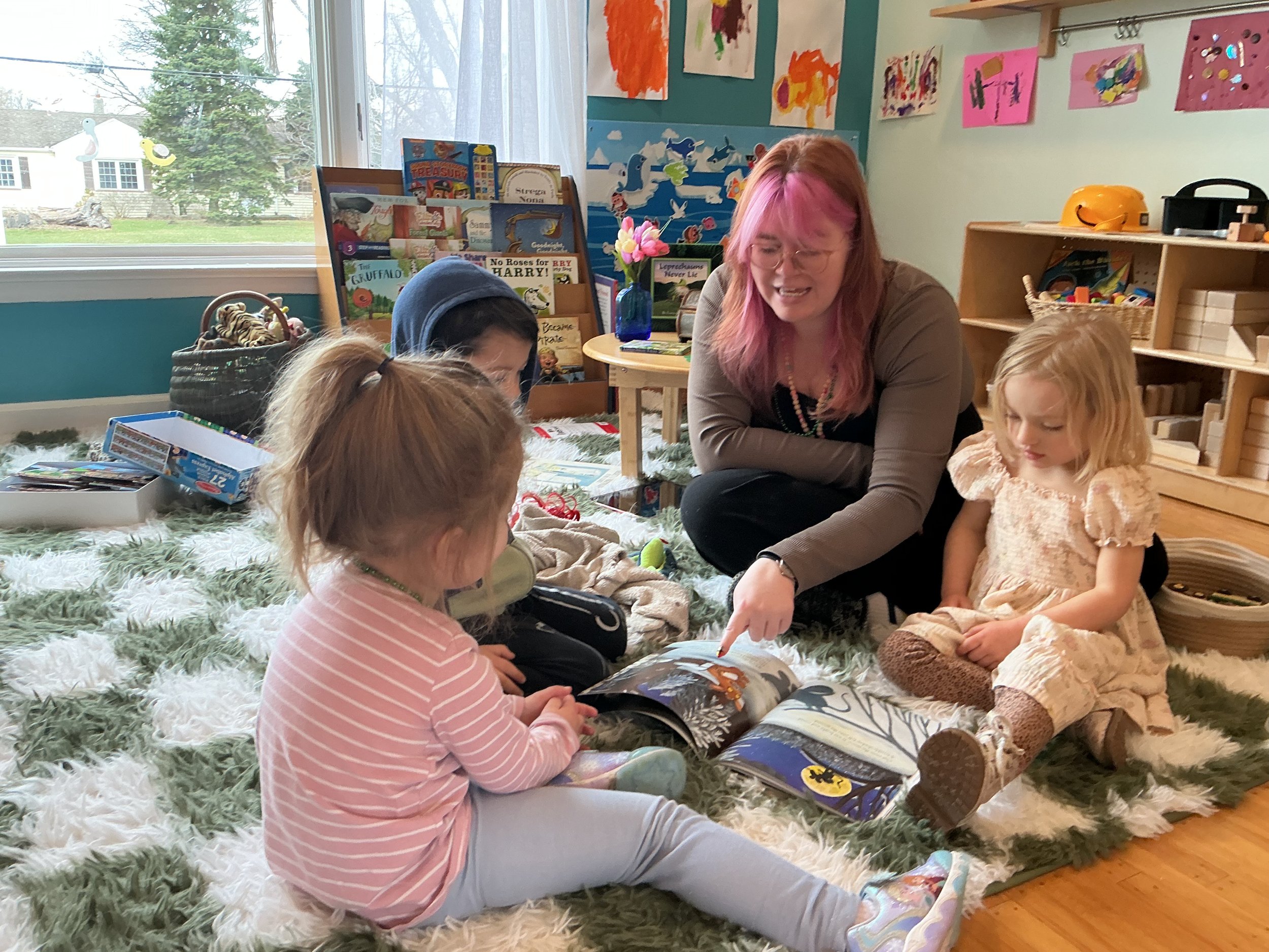 A woman with pink hair and glasses reading a book to three young girls in a colorful classroom, sitting on a fluffy rug.