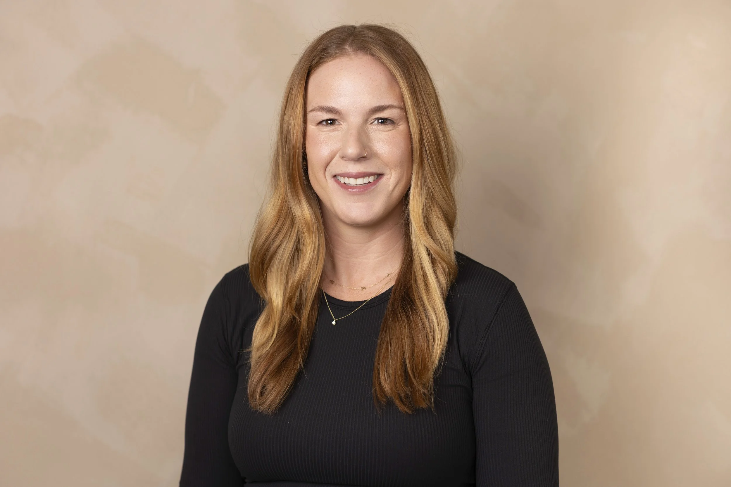 A woman with long red hair smiling, wearing a black top, gold necklaces, and a nose ring, in front of a beige background. Form Modern Pilates, Xformer Pilates, Traverse City MI, Grand Rapids MI