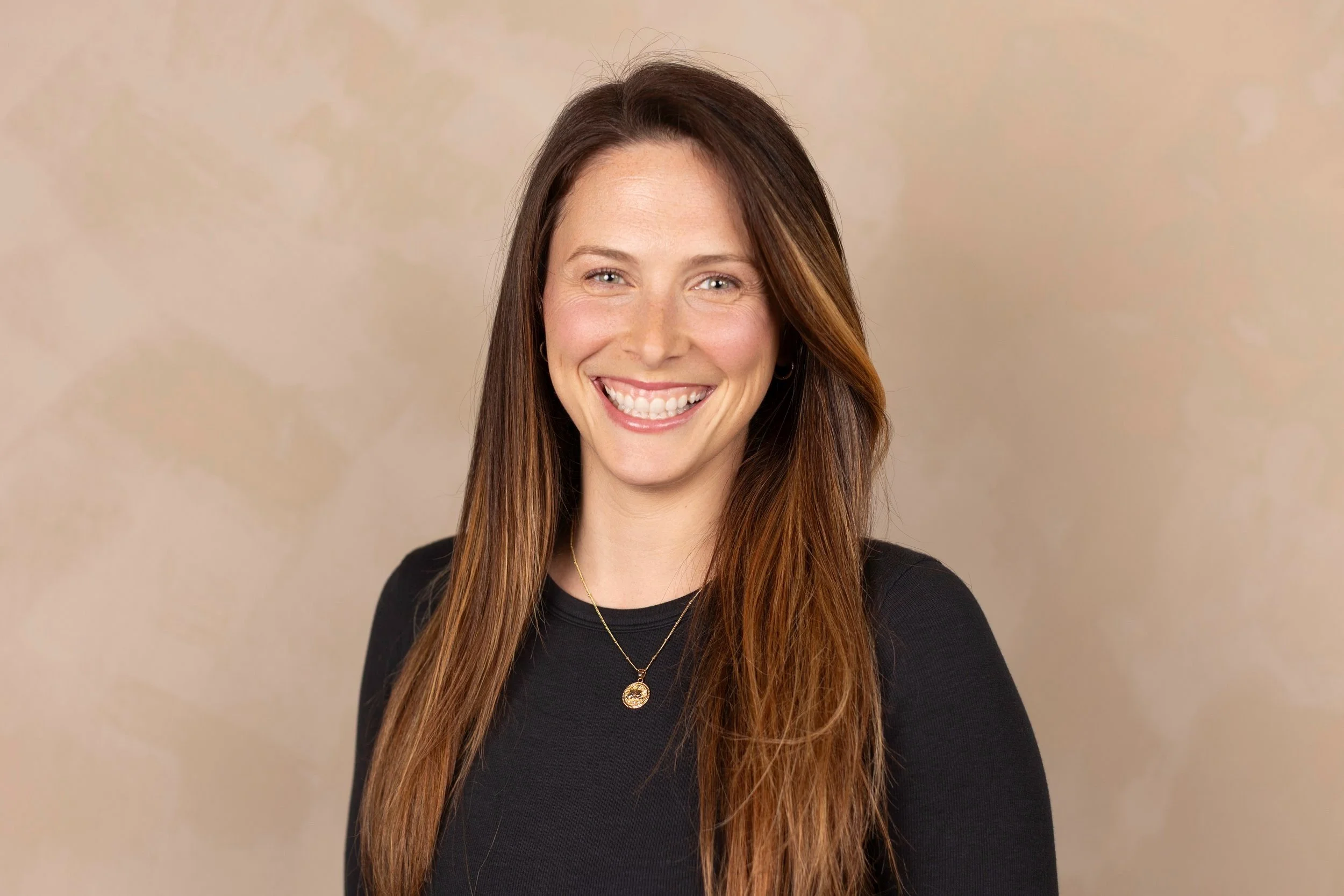 A woman with long brown hair smiling, wearing a black top and a gold necklace, against a beige background.