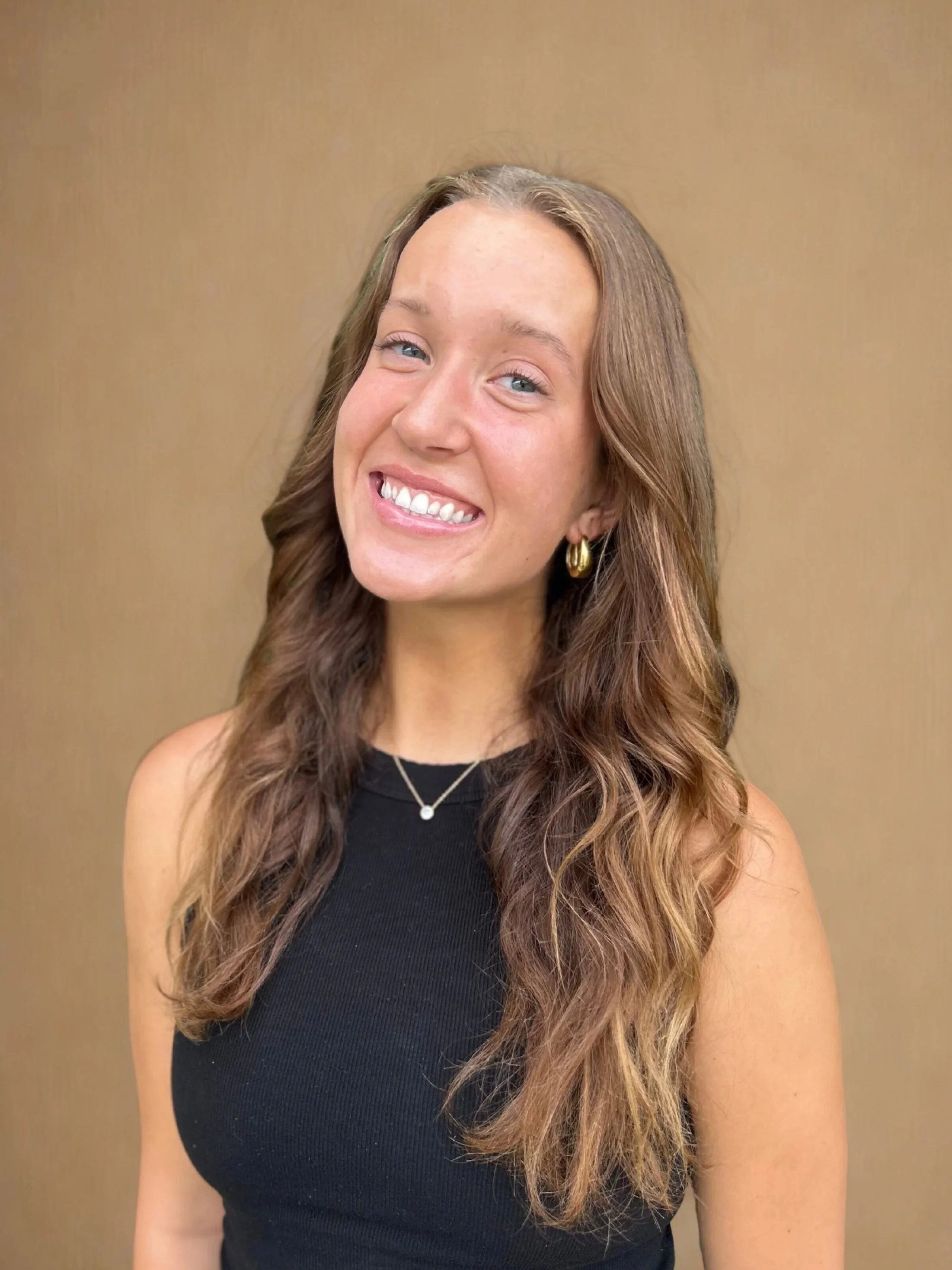 Smiling young woman with long wavy hair, wearing a black sleeveless top, gold hoop earrings, and a delicate necklace, standing against a plain light brown background. Form Modern Pilates, Xformer Pilates, Traverse City MI, Grand Rapids MI