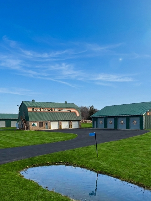 A building with a green roof and a sign reading 'Brad Tanck Plumbing,' located in a grassy area with a curved driveway and a small pond in the foreground.