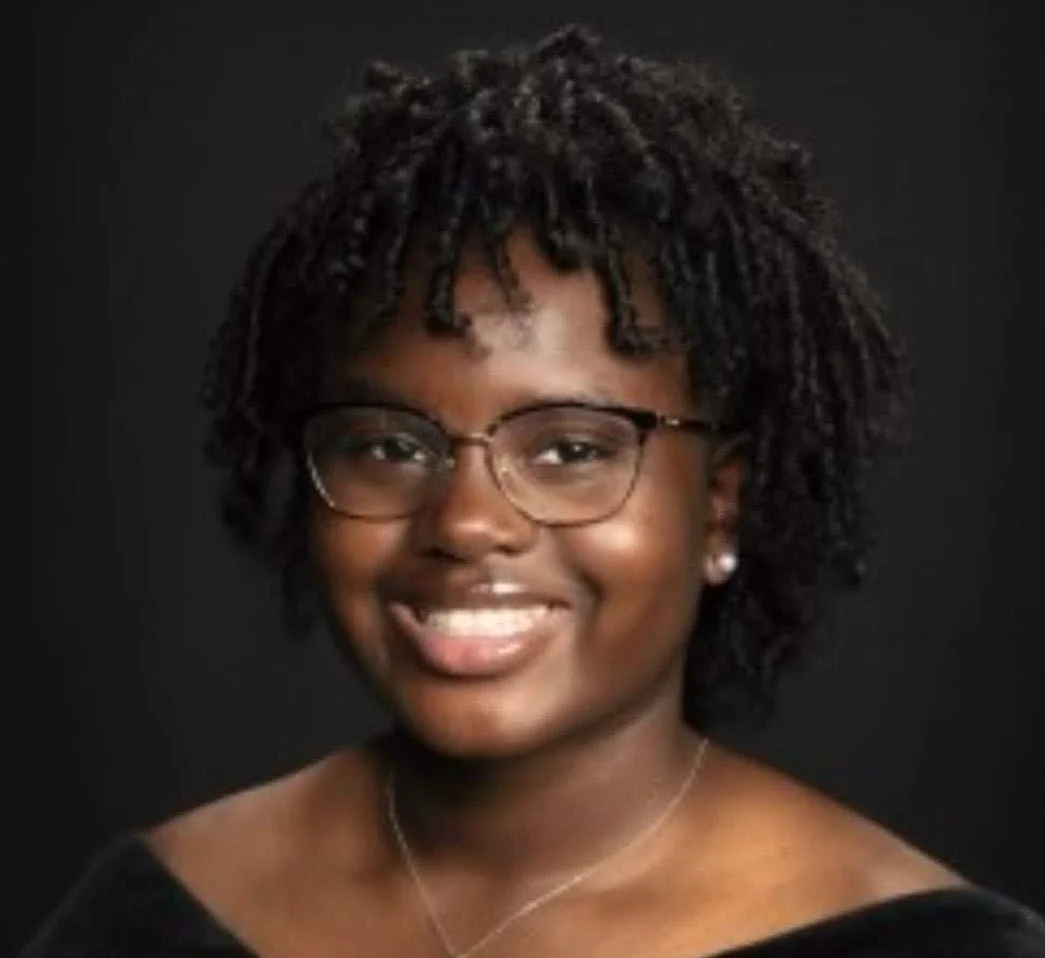 Portrait of a young woman with short curly hair, glasses, and earrings, smiling against a black background.