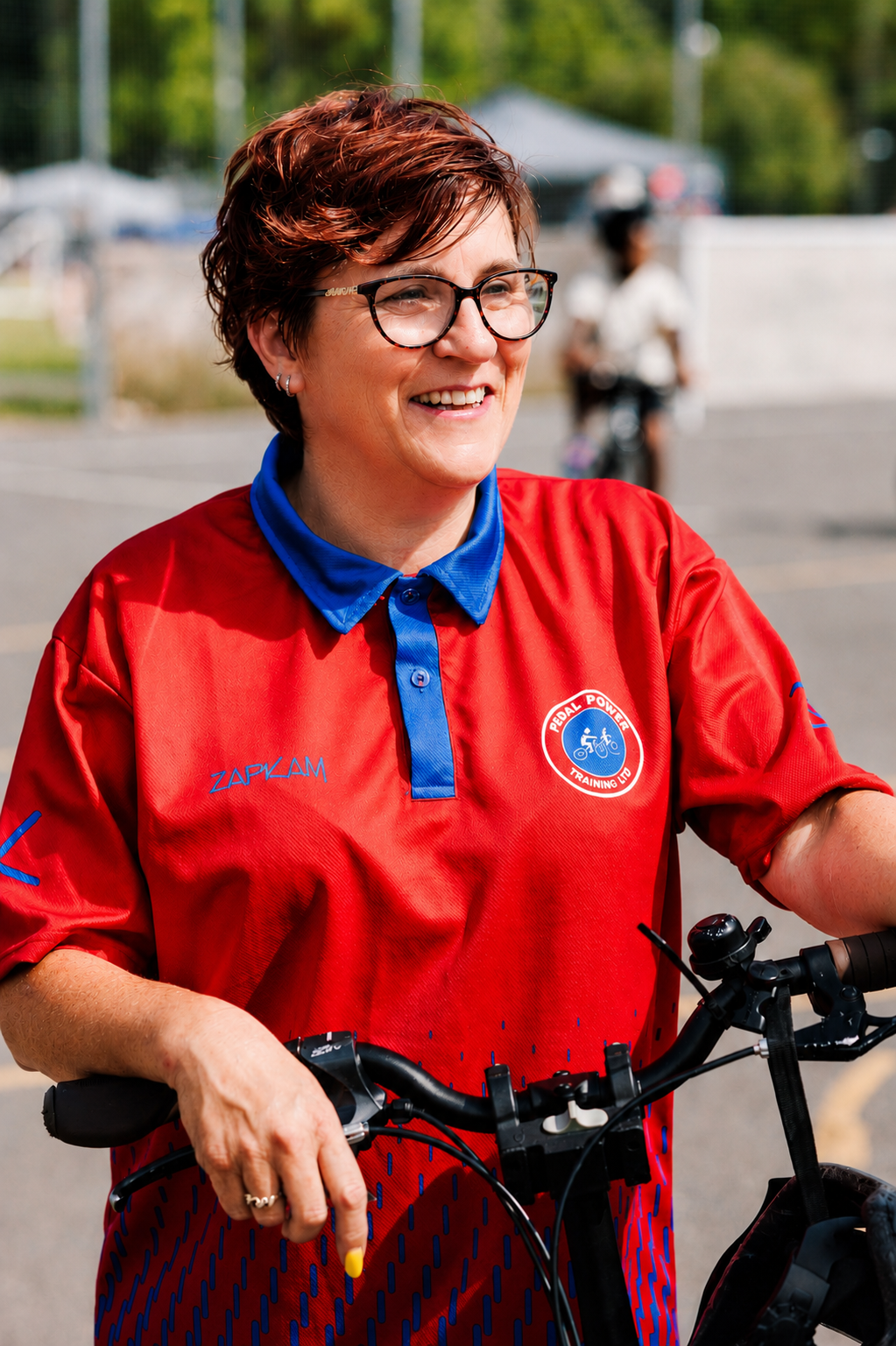 A woman wearing glasses and a red and blue cycling jersey, standing outdoors with a bicycle handlebar in front of her, smiling at the camera.