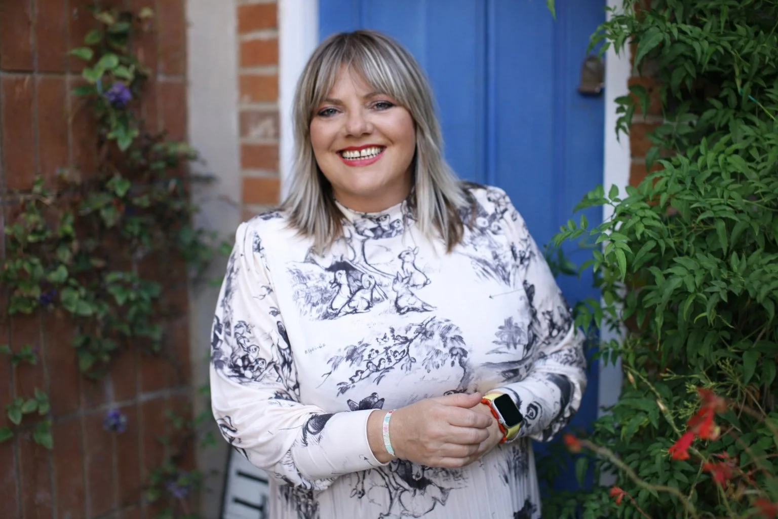 A woman with blonde hair smiling while standing outside near a brick wall and a blue door, surrounded by greenery and plants.
