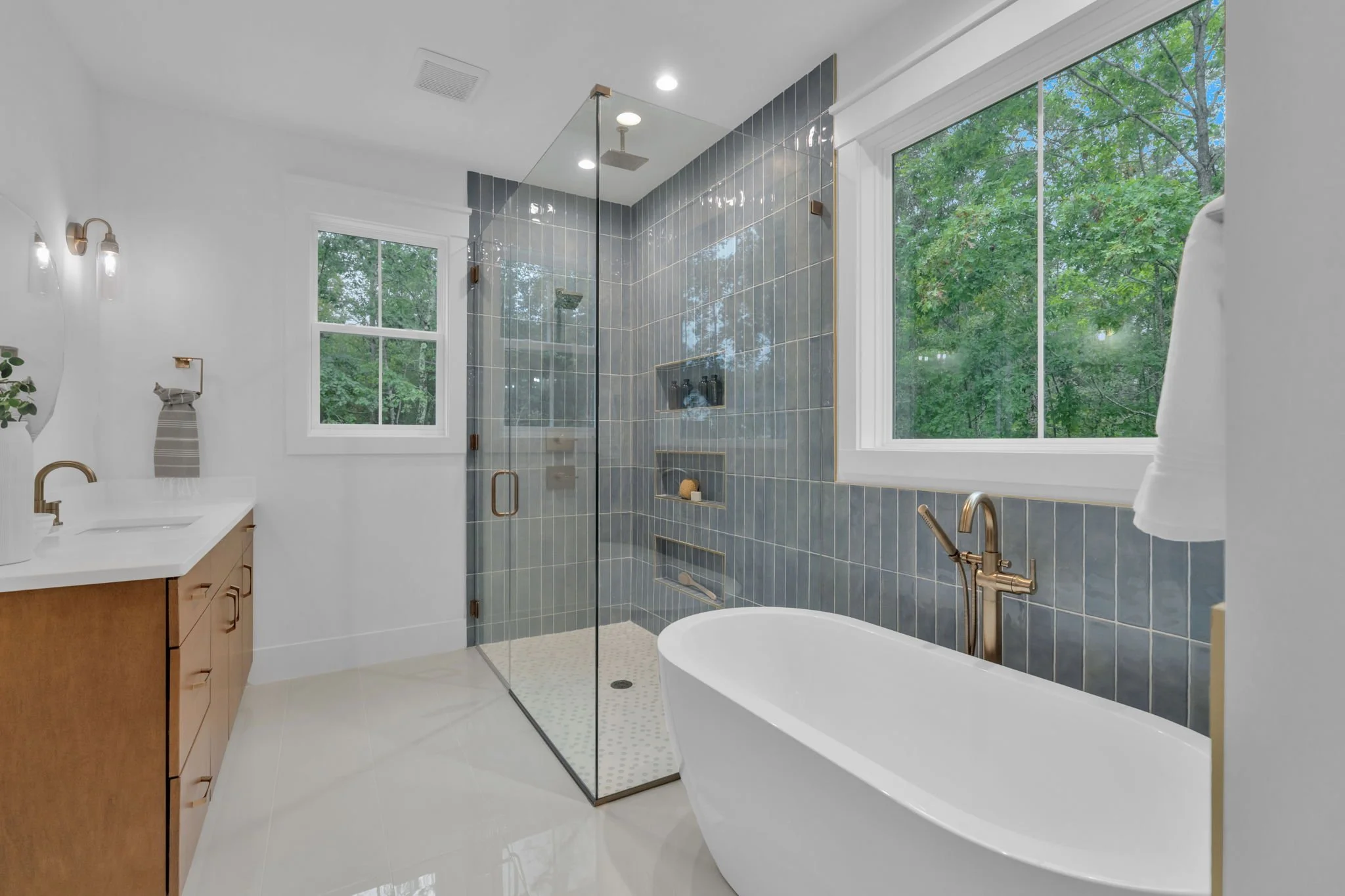 Modern bathroom with a walk-in glass shower, a freestanding bathtub, a wooden vanity with a white countertop, a large round mirror, and a potted plant, featuring light green walls and white trim.