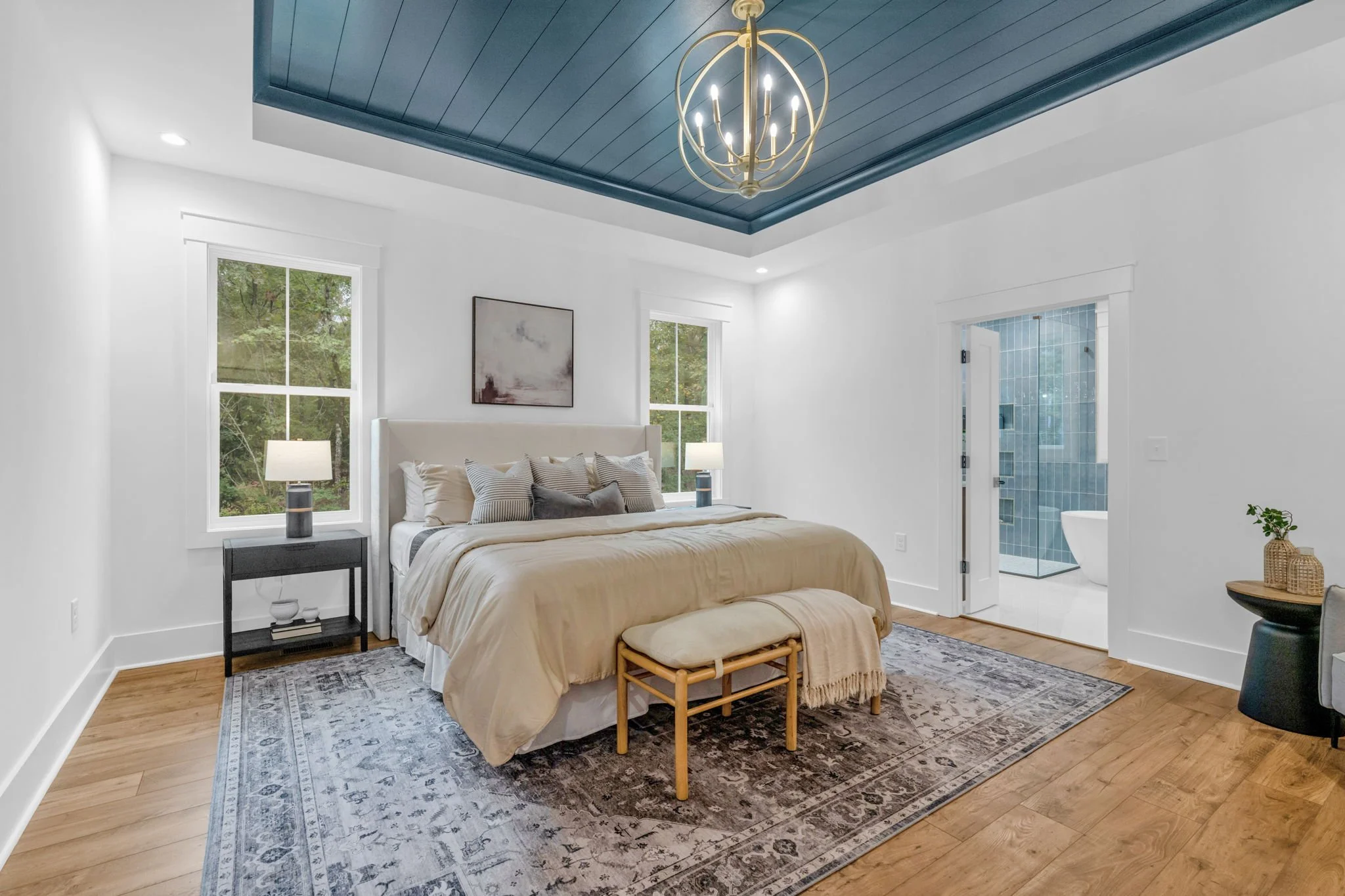 Modern bedroom with beige upholstered headboard, white bedding, and decorative pillows, flanked by two nightstands with lamps, and large windows showing greenery outside. An en suite bathroom with a clawfoot tub is visible through an open door.