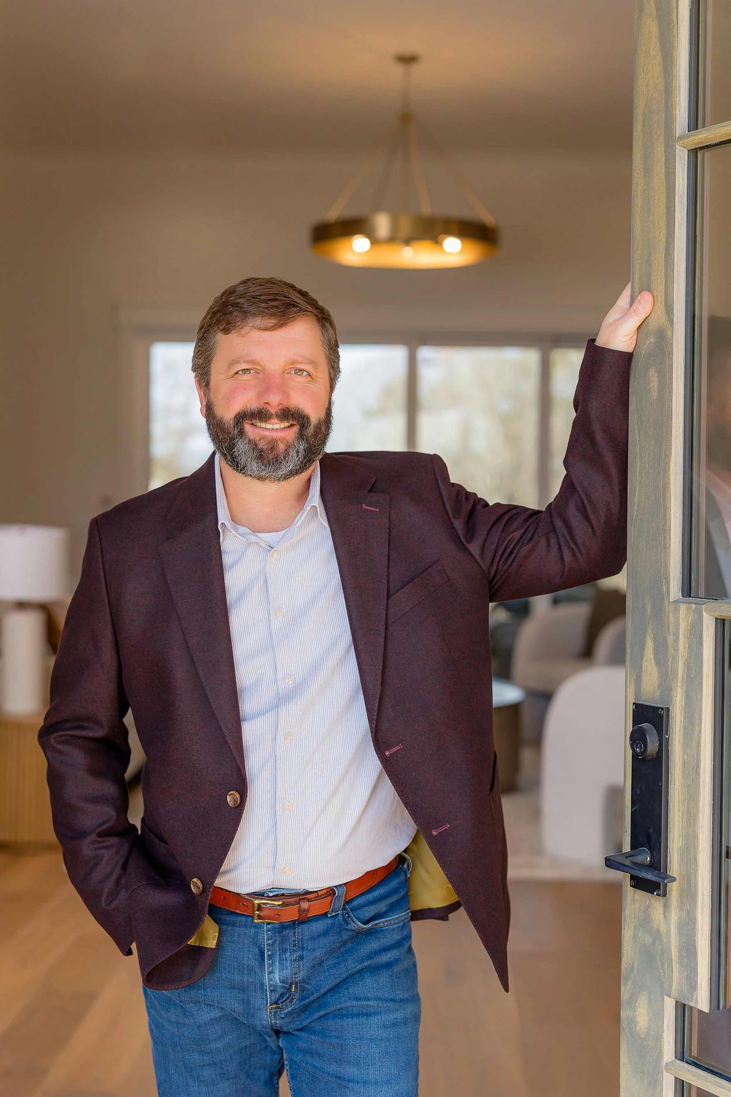 A smiling man with a beard and short hair standing in a bright modern kitchen, dressed in a checkered blazer and white shirt.