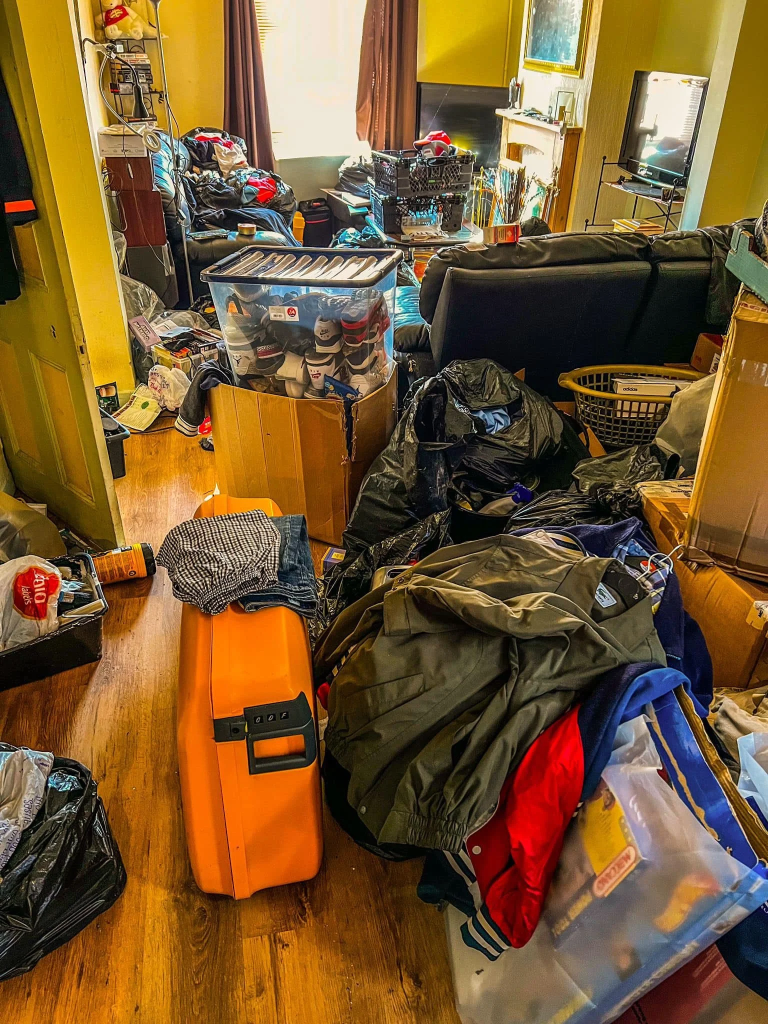 A cluttered living room with piles of clothes, bags, and boxes, including an orange toolbox, and various household items scattered around.