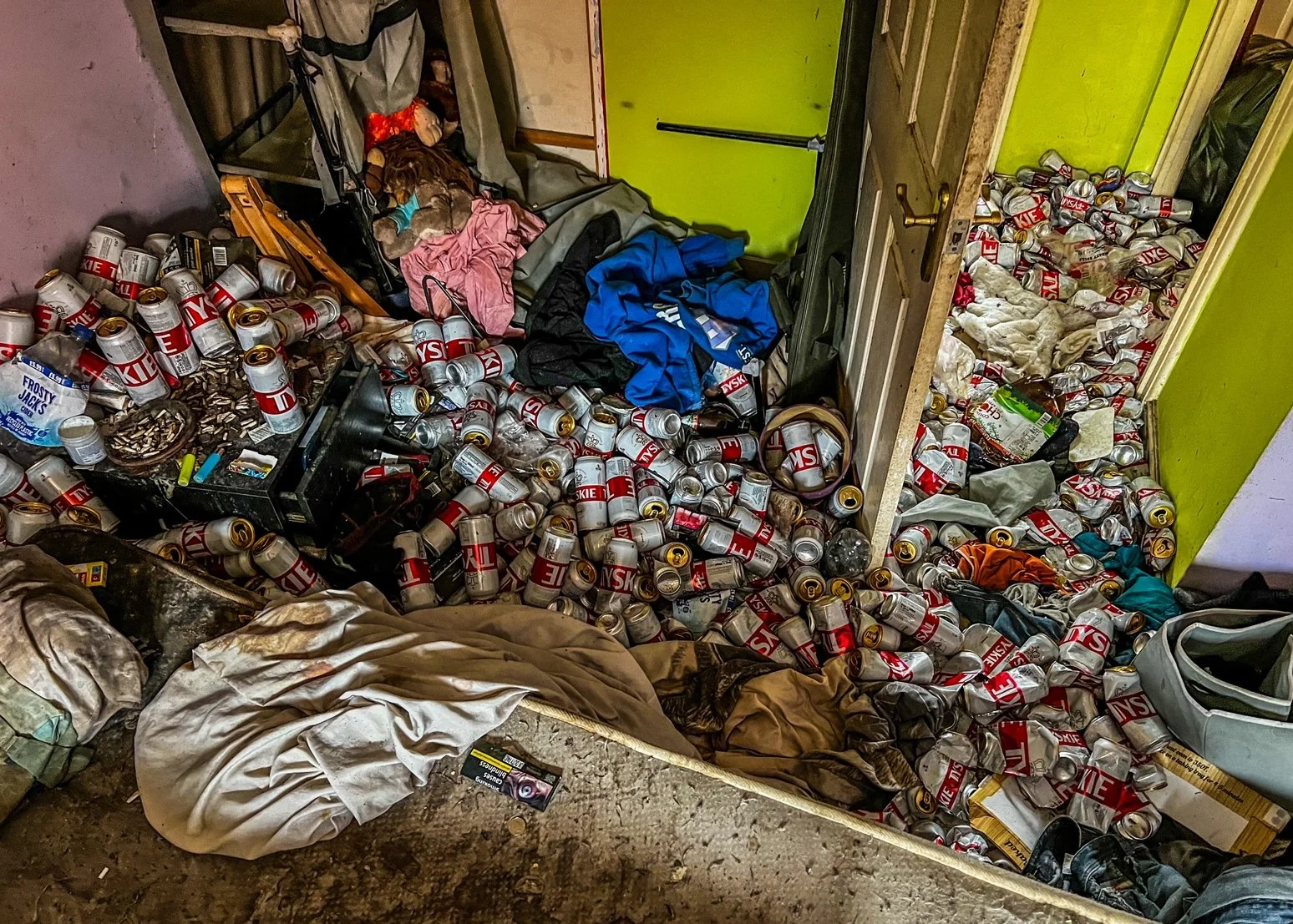 Room filled with empty beer cans, clothes, and trash on the floor.