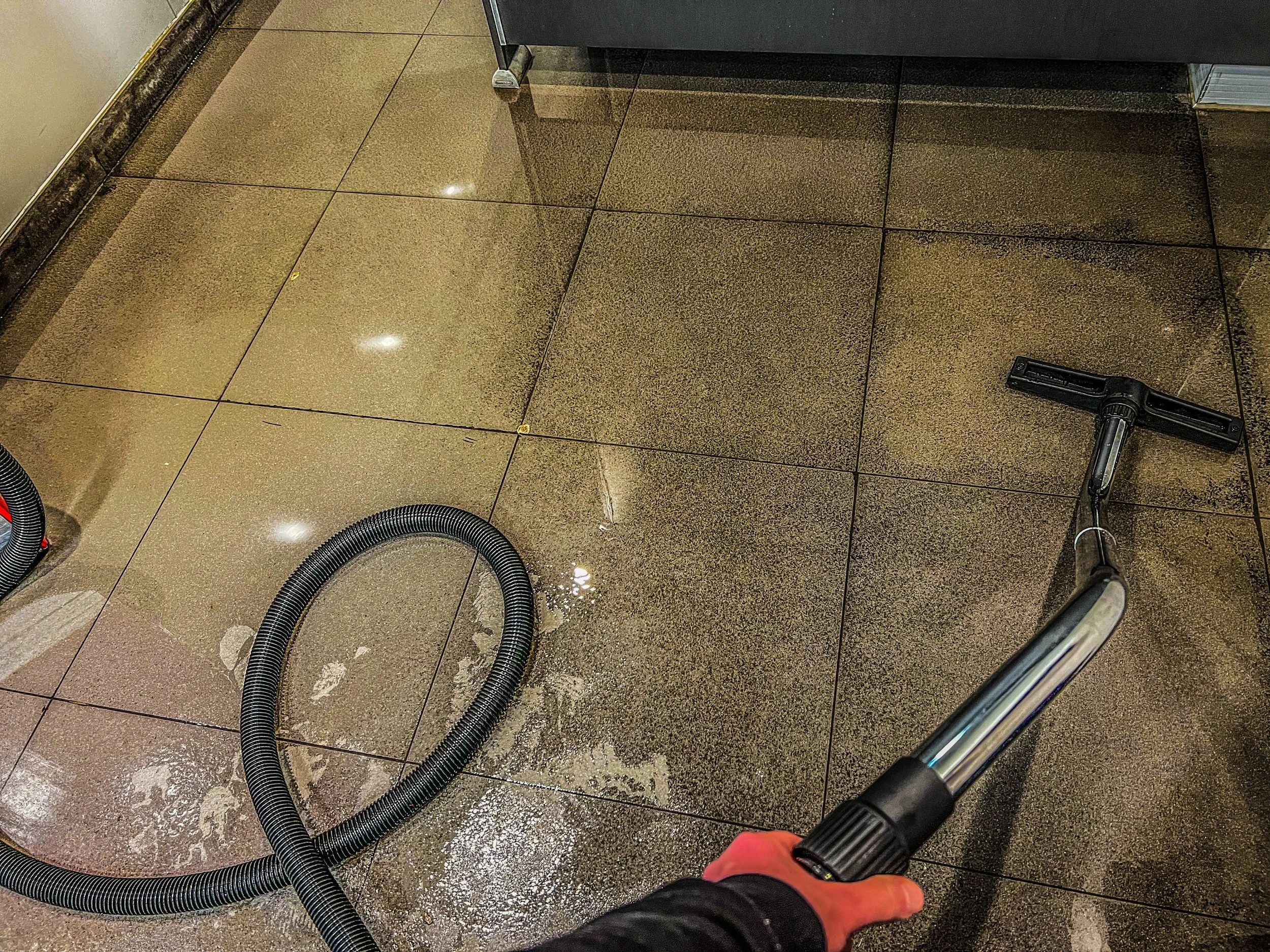 Person using a vacuum cleaner on wet brown tiled floor with hose visible.
