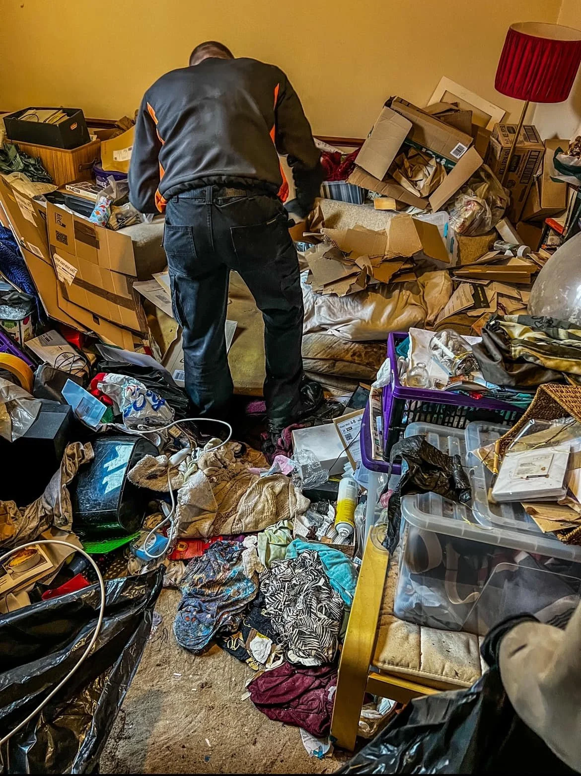 A cluttered room filled with various items, including boxes, clothes, and miscellaneous objects, with a person sorting through the mess.