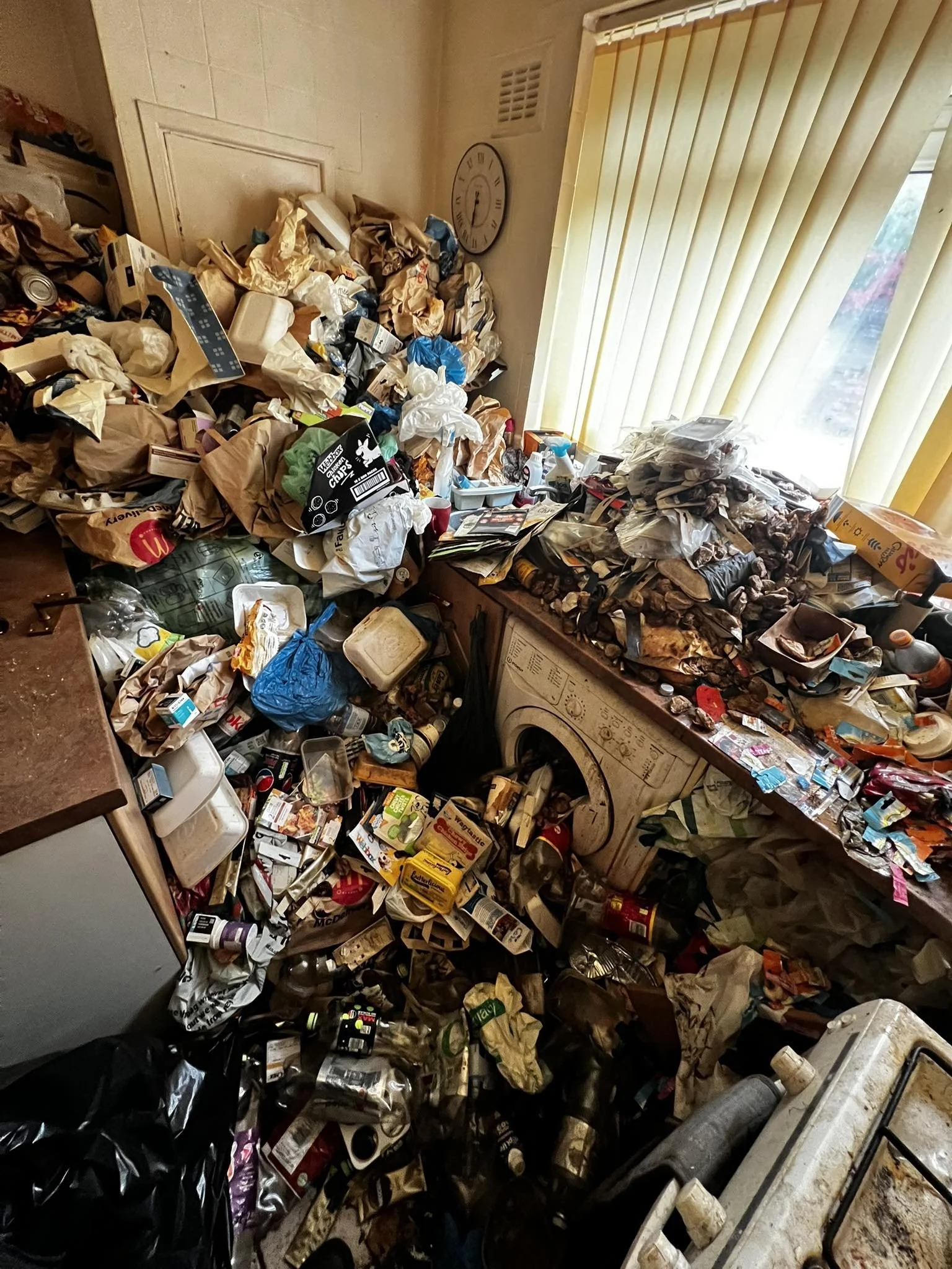 A cluttered laundry room filled with overflowing trash and piled-up items, including paper, cans, and boxes, around a washing machine and near a window with vertical blinds.