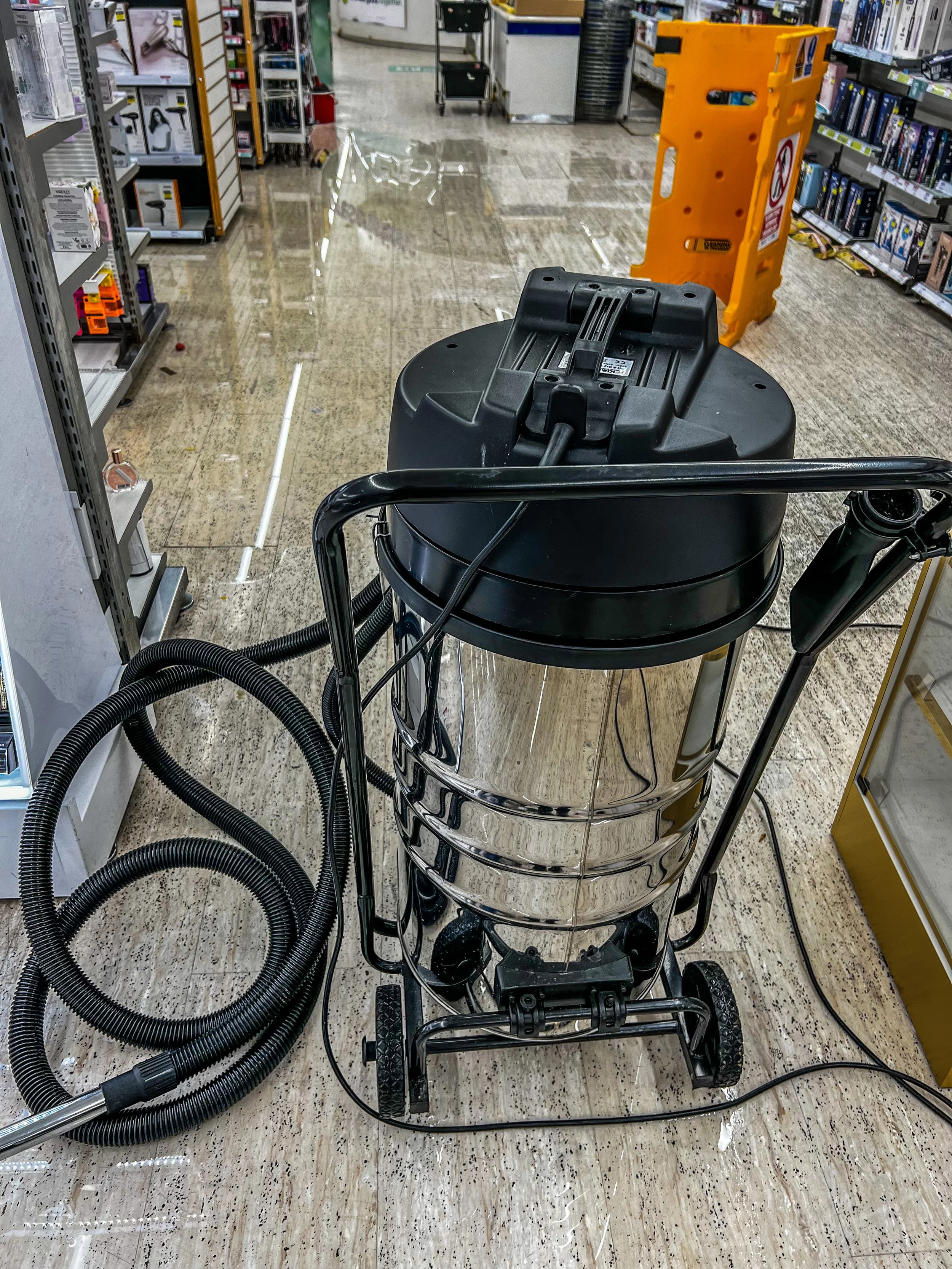 Industrial vacuum cleaner on wet floor in a store aisle, with caution sign in background.