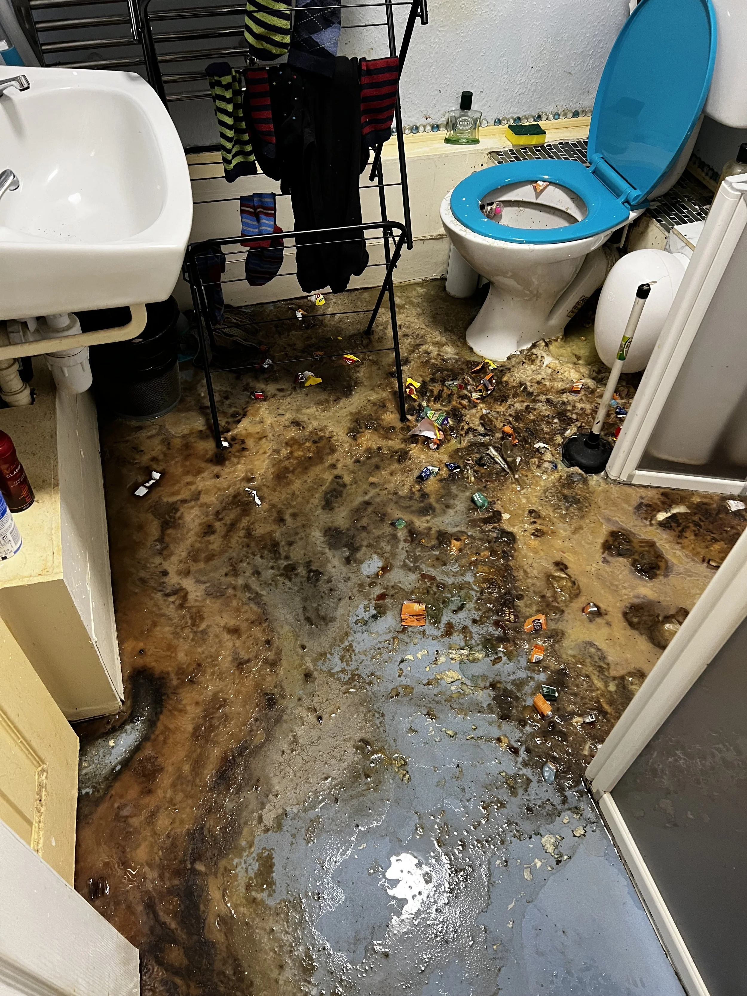 Flooded bathroom with dirty water and spilled trash, including candy wrappers, covering the floor. Visible items include a white sink, a toilet with a blue lid, a mop, a drying rack with wet socks, and various bottles near the toilet.