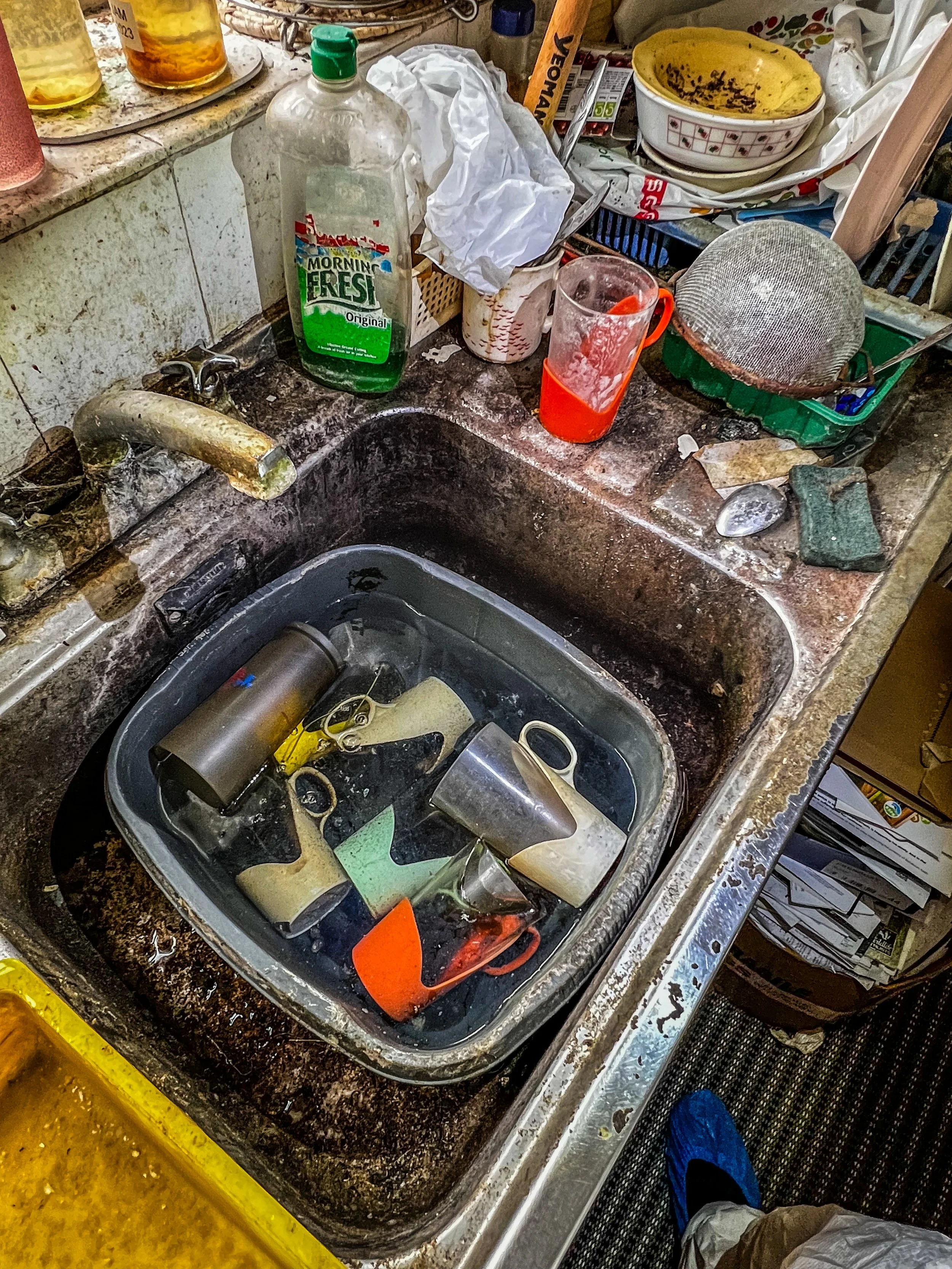 A cluttered kitchen sink with dirty dishes, including mugs and containers, soaking in water. A bottle of dish soap and other items are on the countertop, which appears unclean and disorganized.