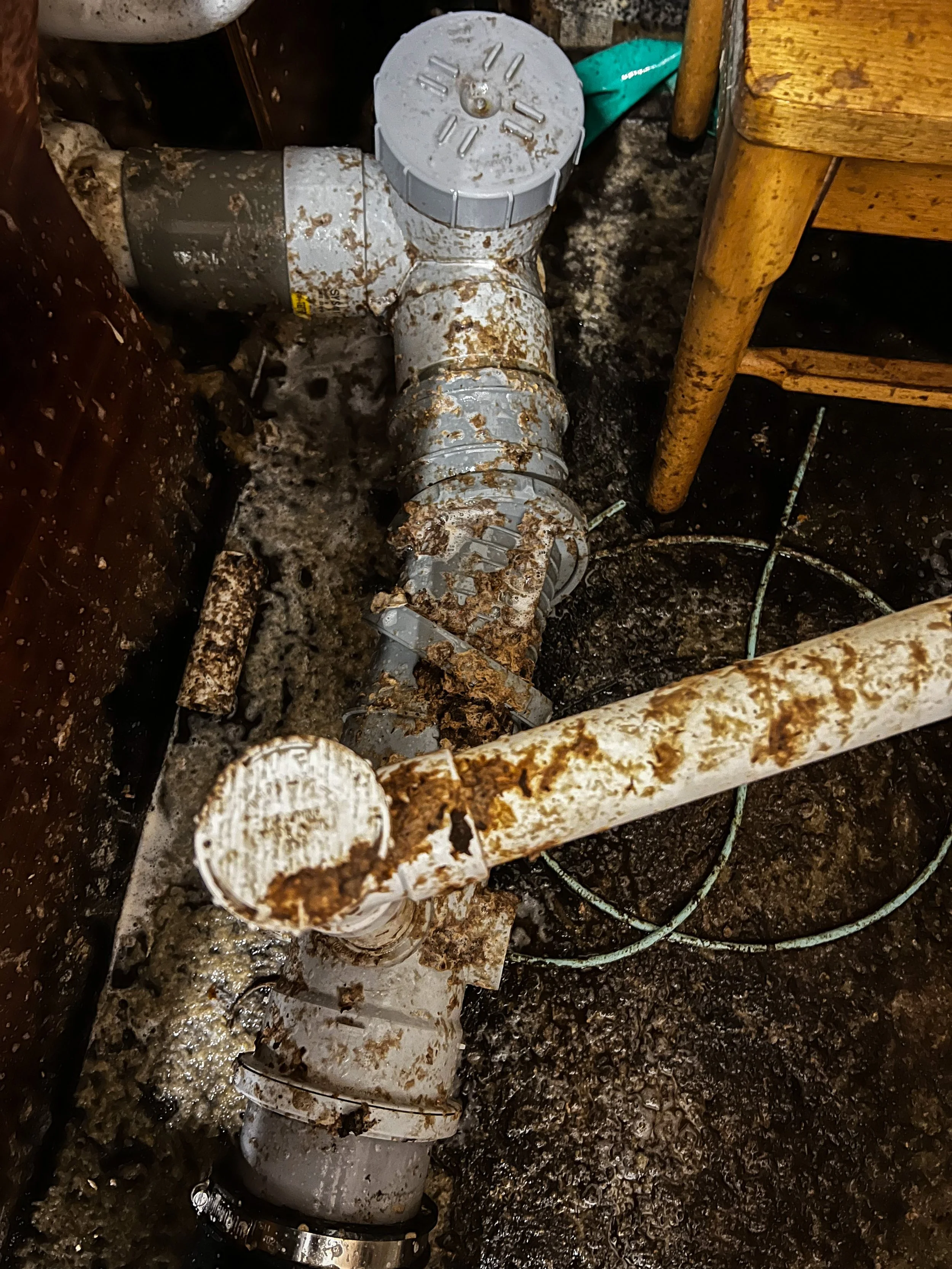 Rusty and corroded plumbing pipes in a damp, cluttered basement with a wooden chair.