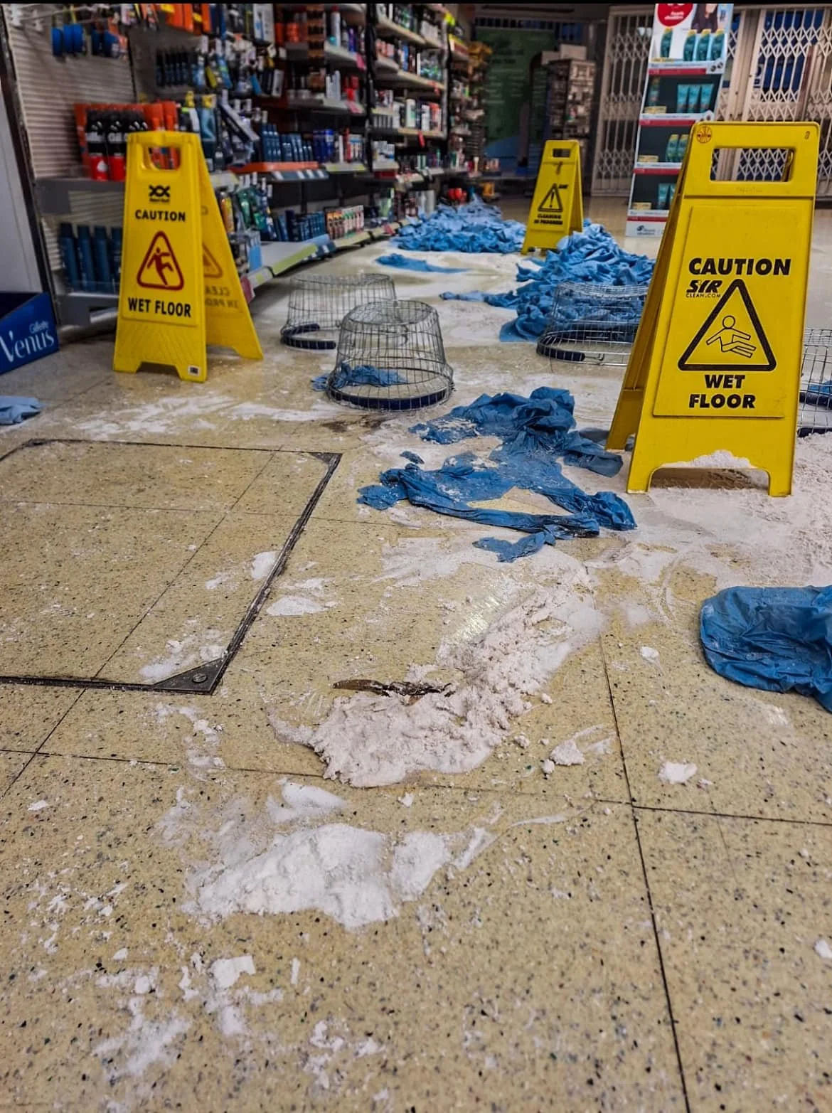 A shopping aisle with wet floor warning signs, scattered blue gloves, and cluttered shelves.
