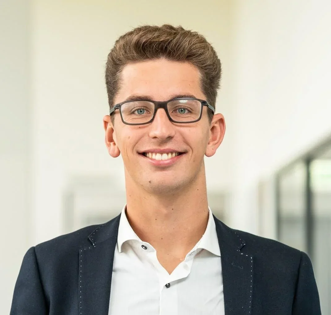 Ein Mann lächelt, trägt eine Brille und ein Jackett.