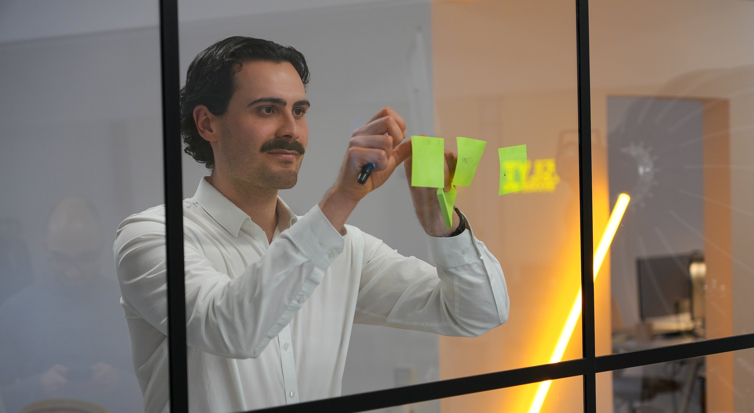 Marco trägt ein weißes Hemd und klebt Post-Its an eine Glaswand