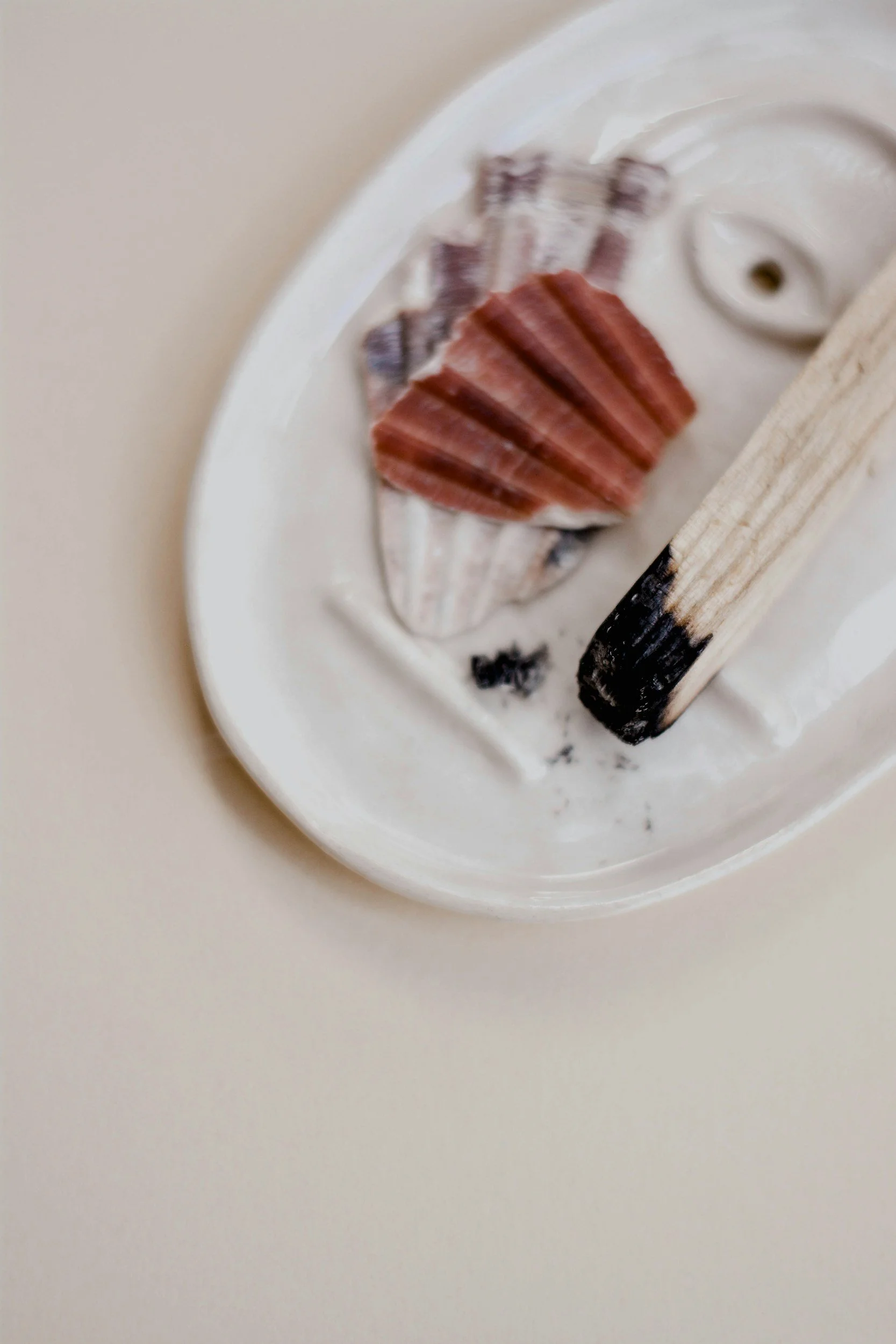 A ceramic dish with a creamy white dip, containing a seashell-shaped snack and a red shell-shaped snack, along with a wooden spoon with a burnt tip.