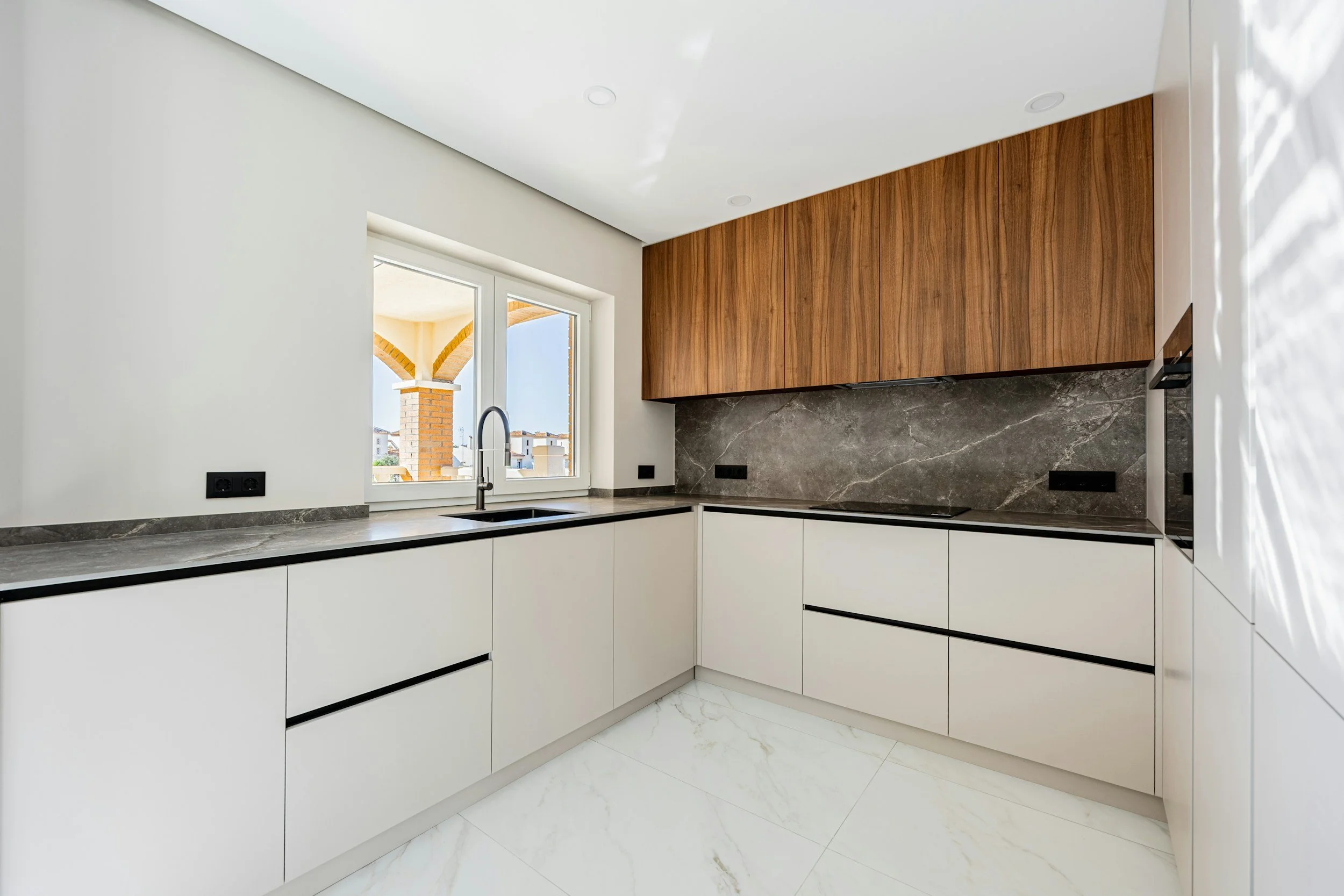 Modern kitchen with white cabinets, black countertops, gray marble backsplash, wooden upper cabinets, and a window showing an outdoor view with brick arches.