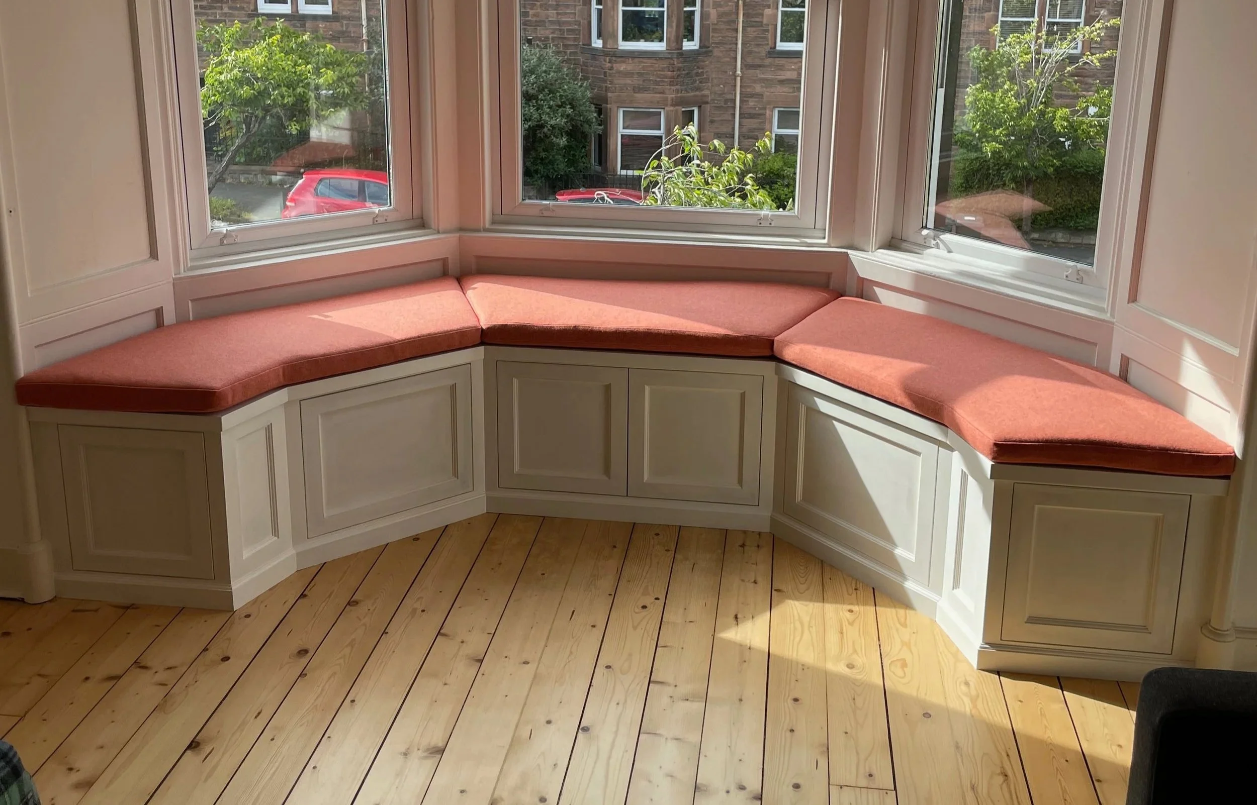 A built-in corner window seat with pink cushions in a bright room, overlooking a garden and brick buildings outside.