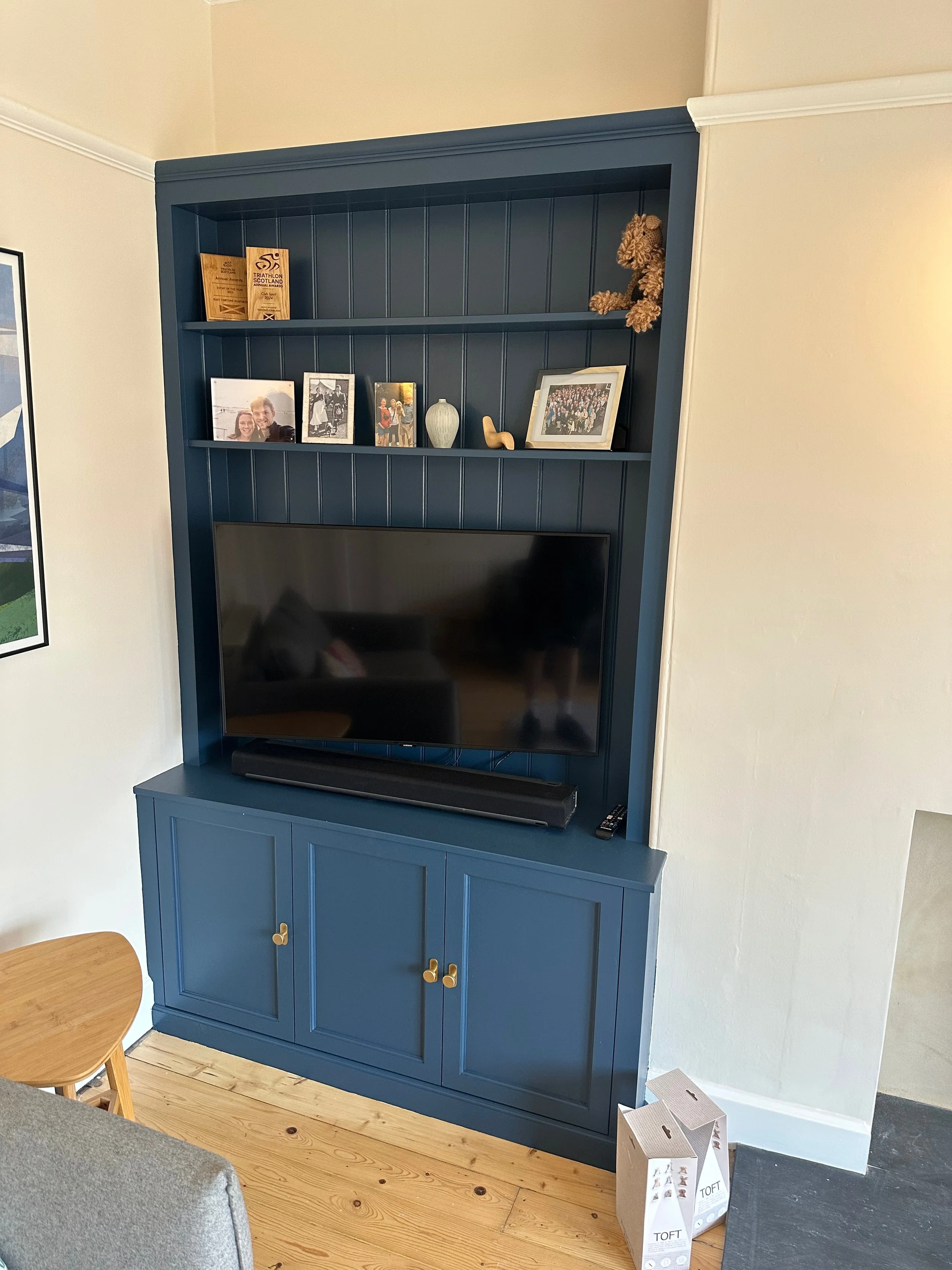 Blue built-in entertainment center with a television, shelves holding photographs, decorative objects, and a teddy bear in a living room.