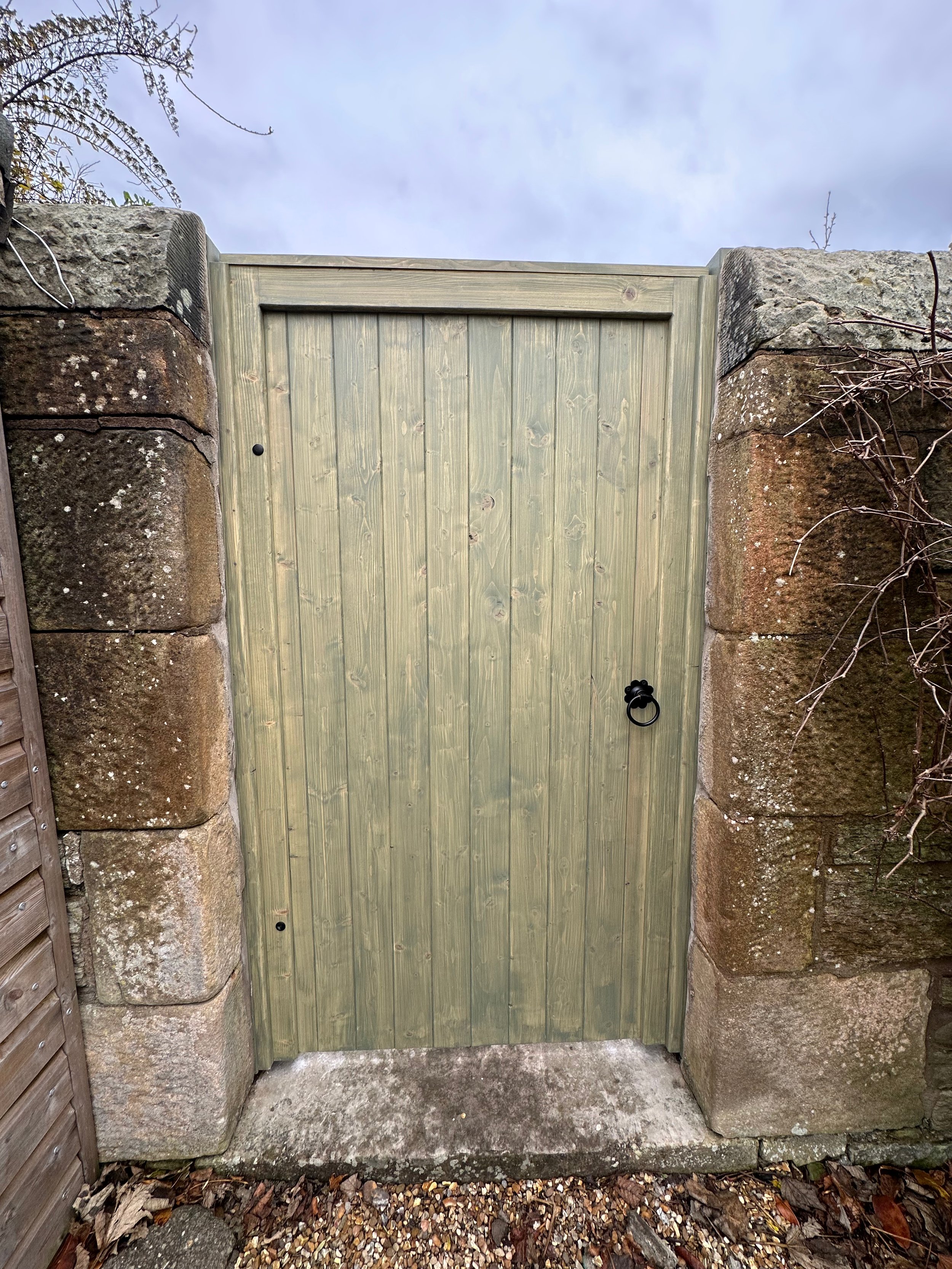 A wooden gate with a black handle set between two stone pillars, with a cloudy sky overhead and leaves on the ground.