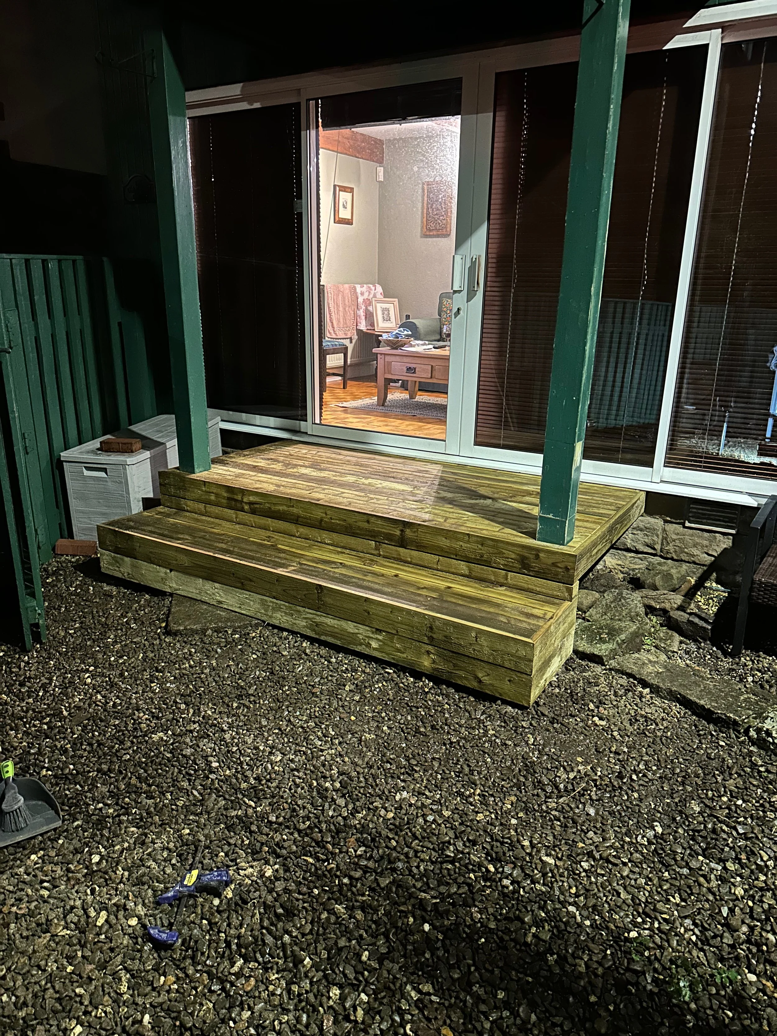 View of a small wooden porch with steps leading up to a sliding glass door, overlooking an interior room with furniture and pictures on the wall. The porch has gravel ground and building supports painted green.