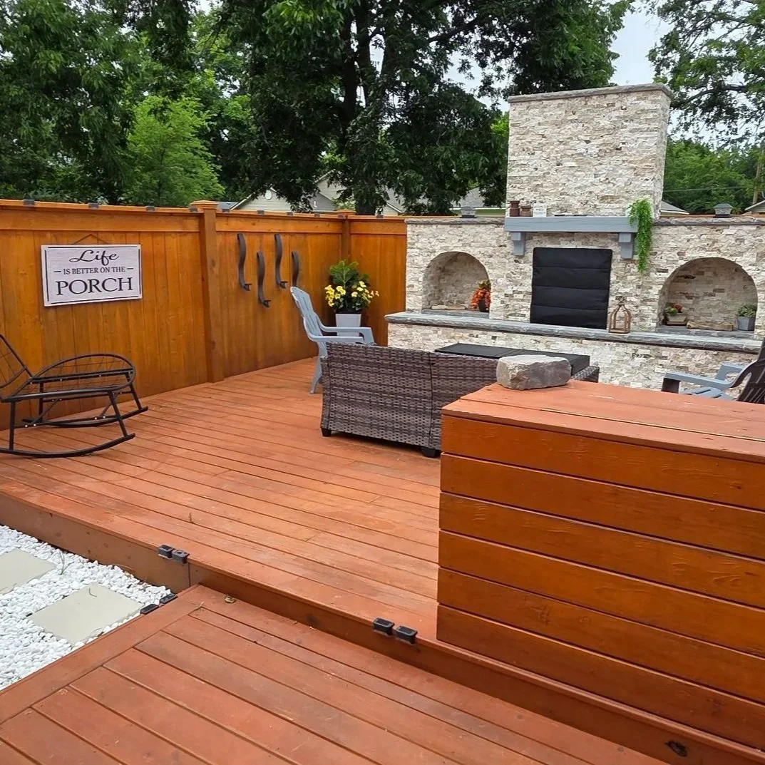 A cozy backyard patio with a wooden deck, outdoor chairs, a stone fireplace, and a decorative sign on the fence that reads 'Life is better on the porch.'