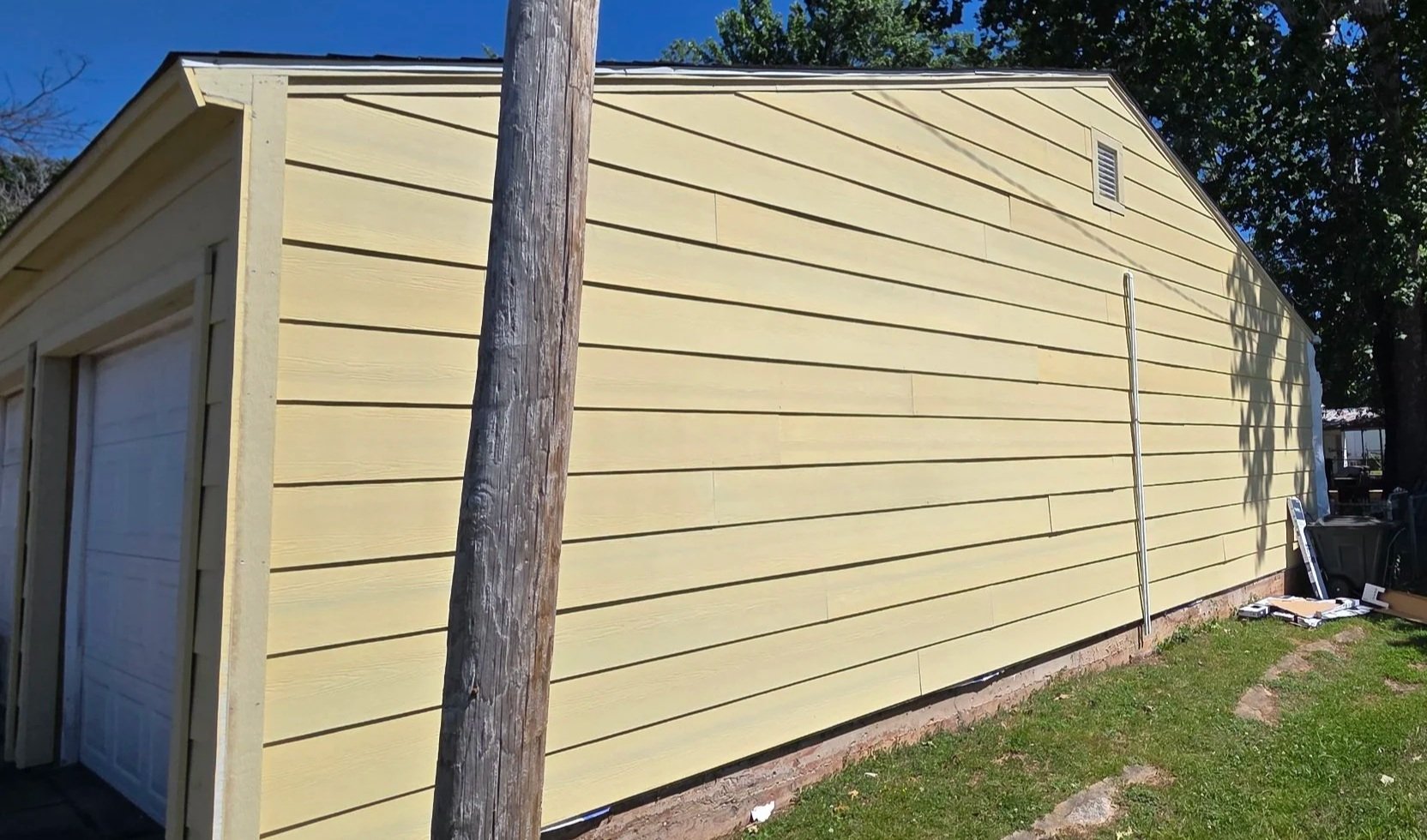 Side of a yellow building with horizontal siding, a small vent near the roof, and a metal pole leaning against it, with tools and equipment on the ground nearby.
