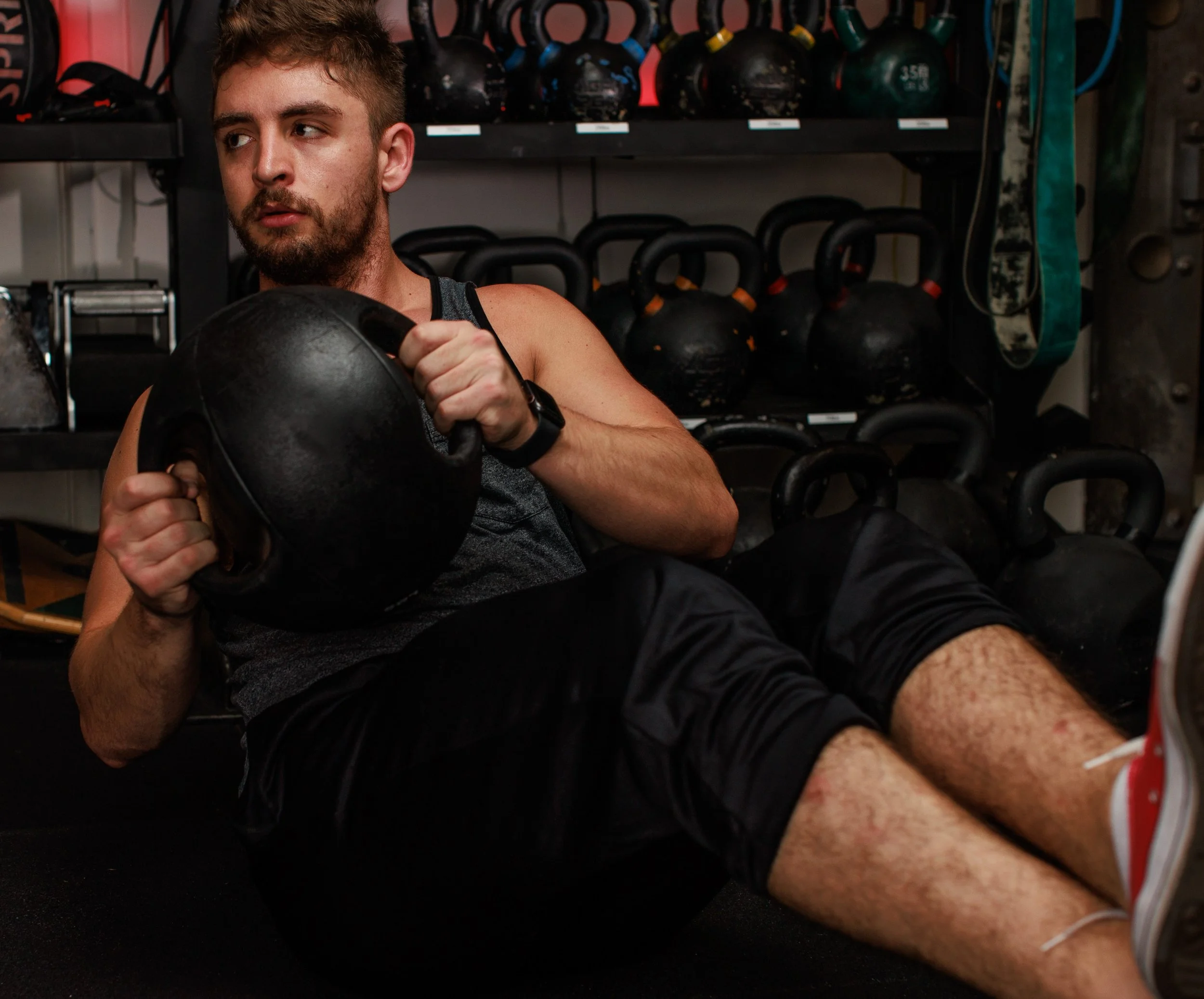 Coach Cooz sitting on the floor in a gym, holding a medicine ball with both hands, surrounded by kettlebells on shelves behind him.