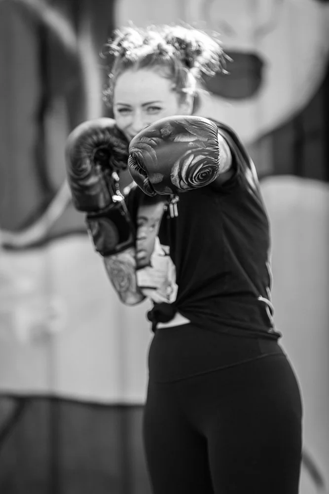 A woman in athletic clothing and boxing gloves, smiling and throwing a punch during a training session, in black and white.