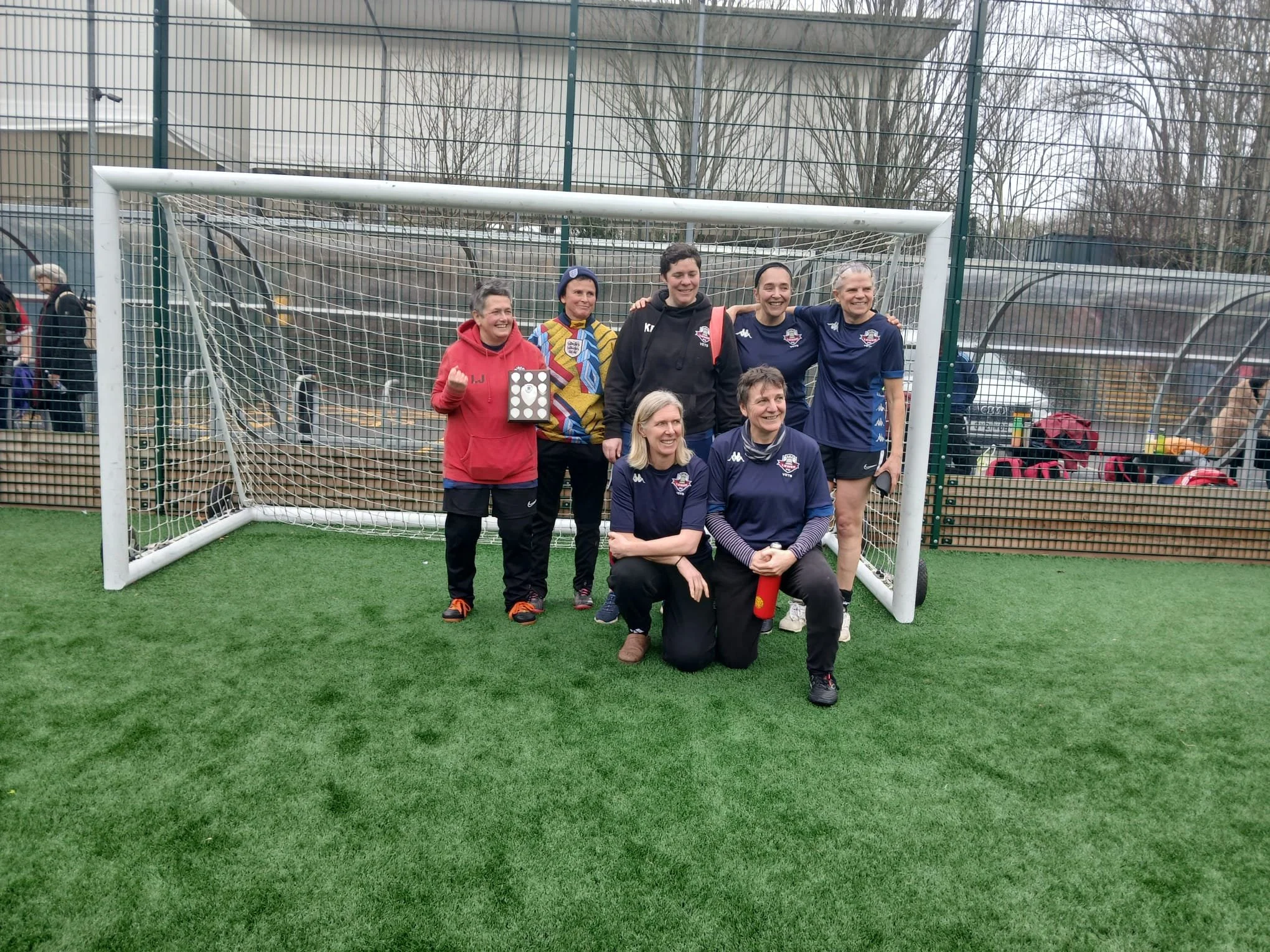 Group of seven women standing and kneeling in front of a soccer goal on an artificial turf field, smiling and posing for a photo, with some women wearing navy blue sports uniforms and two women in casual sports clothing.