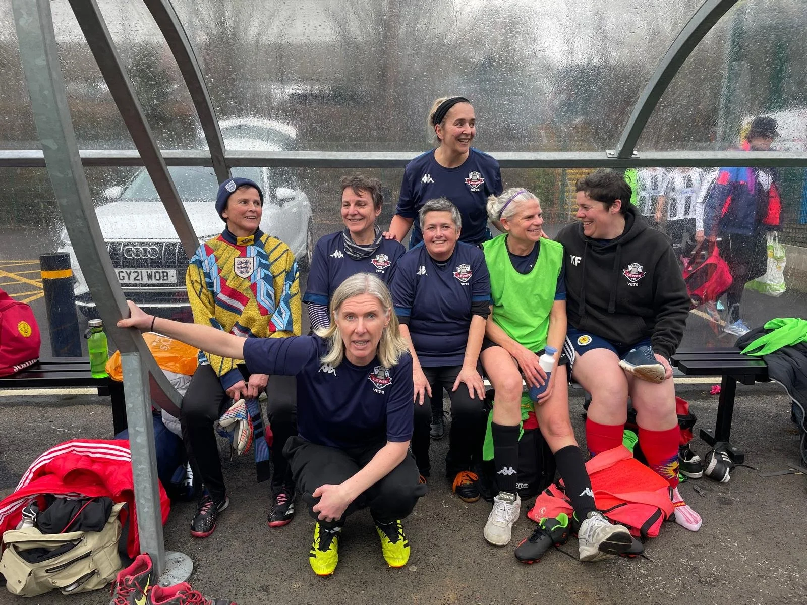 Women’s soccer team sitting on a bench under a rain-soaked shelter, smiling and celebrating after a match, with sports gear around them.