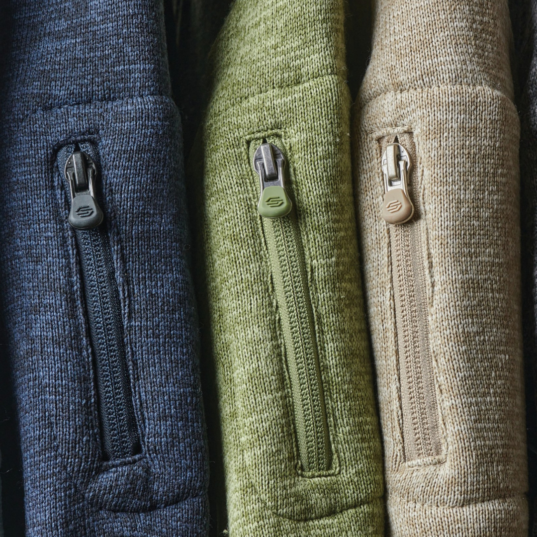A pair of black rubber rain boots with gray and black striped socks draped over the top, placed on a concrete garage floor with a black toolbox and yellow container in the background.